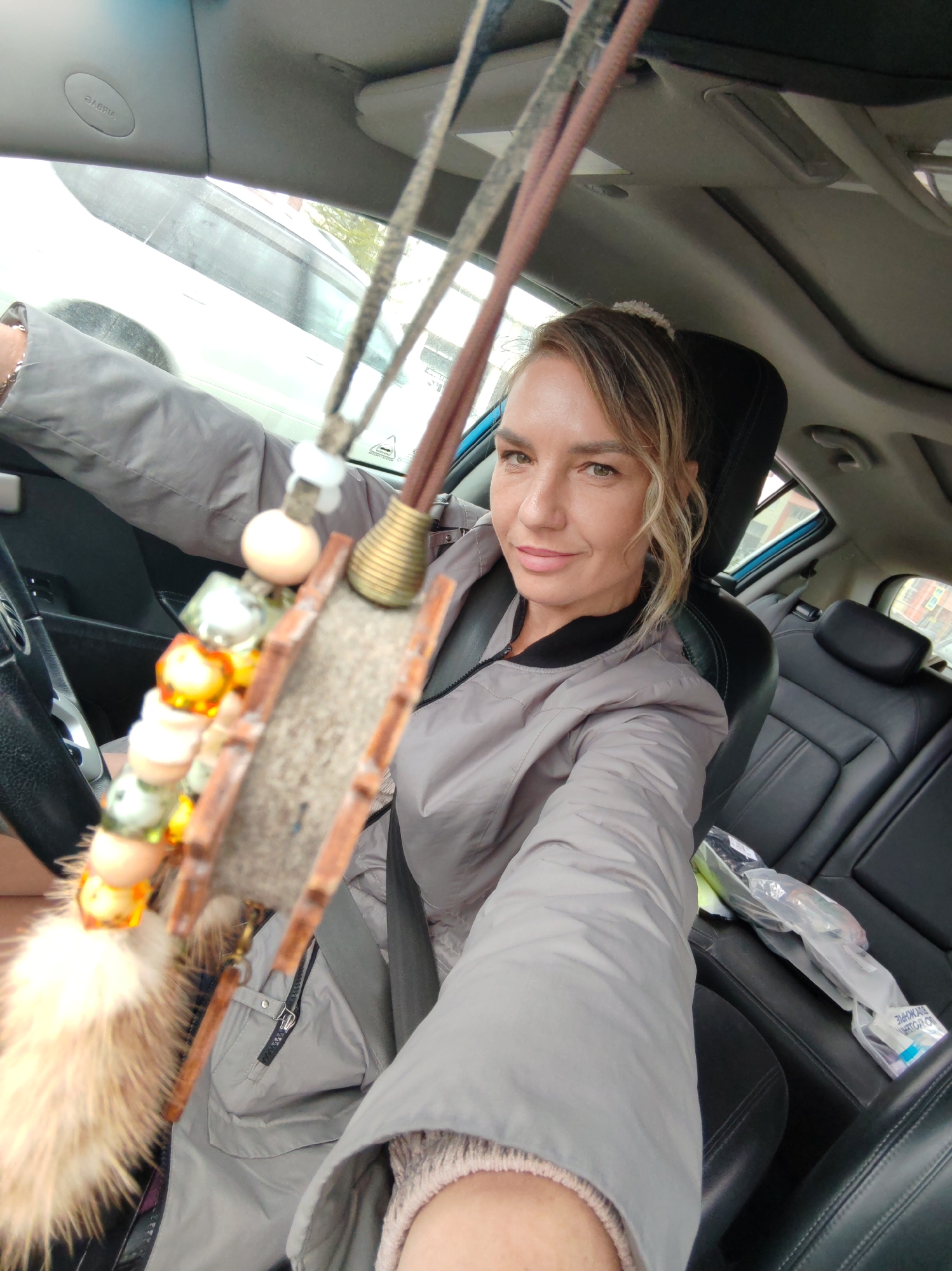 A blonde woman is sitting in the driver's seat of a car, looking at the camera. She is wearing a gray jacket. In the foreground, there is a car accessory hanging from the rearview mirror, which is blurred. The interior of the car is visible, including the dashboard, steering wheel, and seats. The woman appears to be in Petropavlovsk-Kamchatsky, Russia, as suggested by context clues from the user's prompt.
