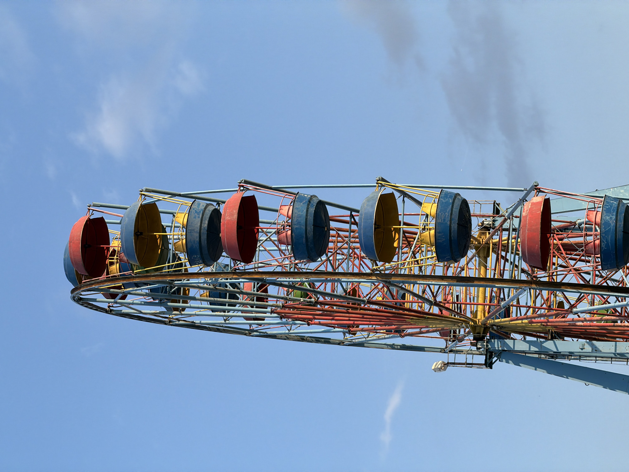 A close-up, low-angle shot captures the side of a colorful, aging Ferris wheel against a clear blue sky with scattered wispy clouds. The Ferris wheel features red, blue, and yellow gondolas, each with a red top, blue base, and yellow interior. The metallic structure of the wheel is painted in red and blue, showing signs of wear and rust. The gondolas are suspended from the wheel's rim by thin, red metal supports. The left side of the image shows a large blue support beam and a small platform with a light fixture attached to it. The scene appears to be set in an amusement park or fairground, though no other structures or people are visible.