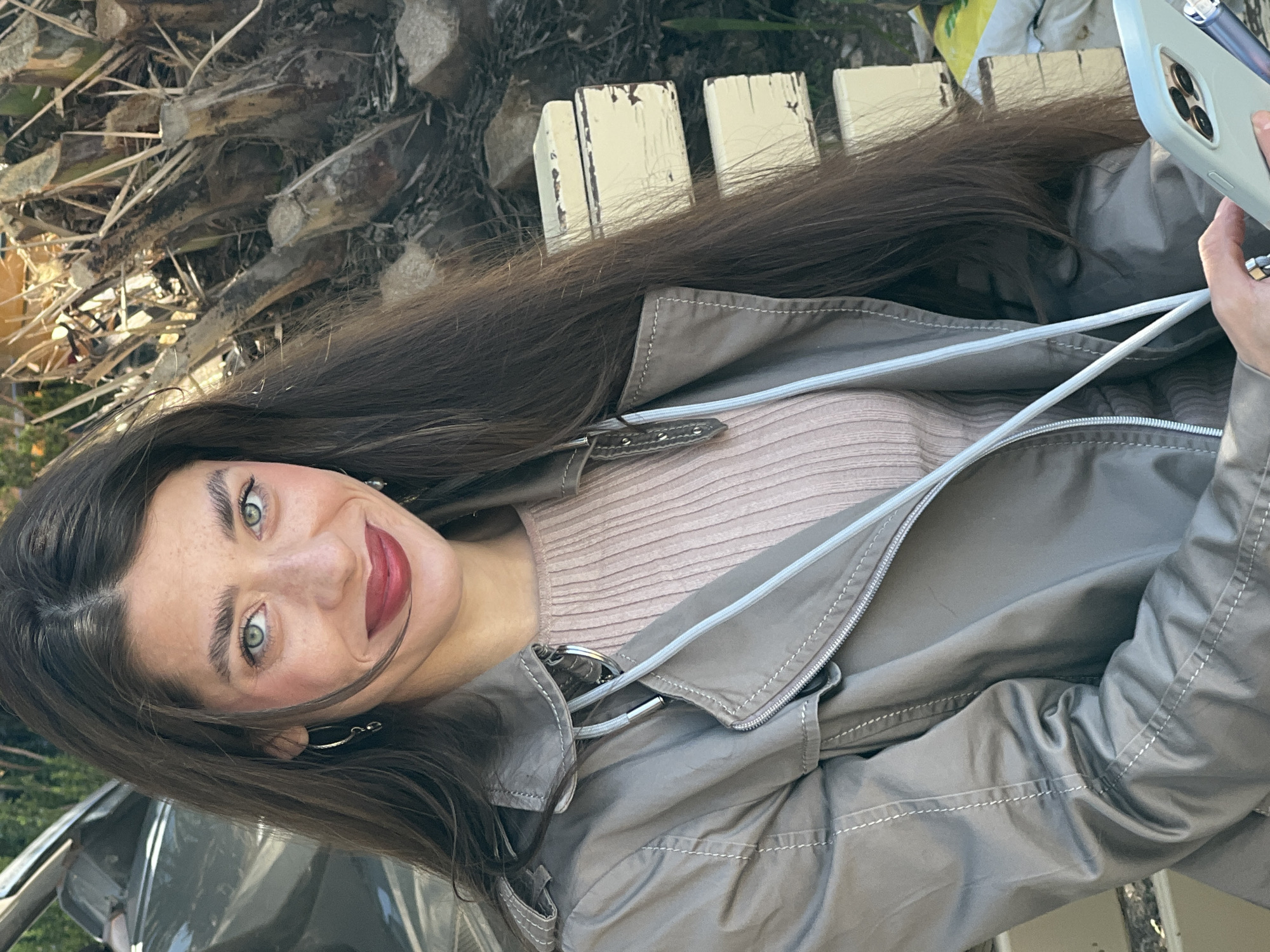 A woman with long, dark hair and green eyes is taking a selfie with a smartphone outdoors. She is wearing a light pink ribbed top and a gray, zip-up jacket. A palm tree trunk and weathered wooden fence posts are visible in the background, suggesting an outdoor location possibly near a park or public space. The exact location within the city of Budva, Montenegro, cannot be determined from the visual information alone.