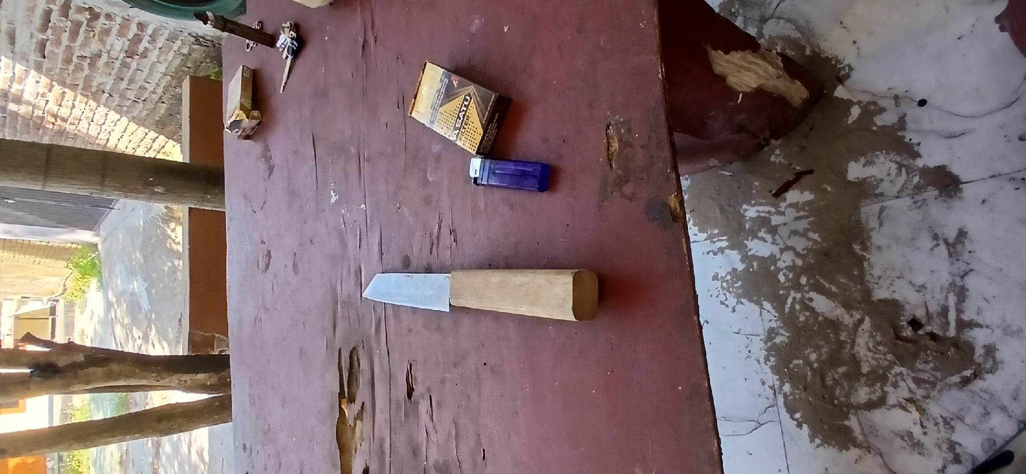 The image depicts a close-up, overhead view of a weathered, dark red wooden table outdoors. A single, silver-bladed knife with a light-colored wooden handle is positioned vertically on the left side of the table. To the right of the knife, there is a small, blue plastic lighter, and further to the right, a partially visible pack of cigarettes, with the brand name "Dji Sam Soe FILTER" visible. In the upper right corner, a set of keys with a keyring is visible, along with a small, rectangular object that appears to be a lighter or a similar item. The table's surface shows signs of age and wear, with visible cracks and indentations. The background reveals a brick wall bathed in sunlight, casting long shadows. Trees are visible in the upper left portion of the image, and the ground beneath the table appears to be concrete with scattered debris and stains. The scene is likely set in a casual outdoor area, possibly a yard or patio, in Pejaten, Indonesia, given the context.