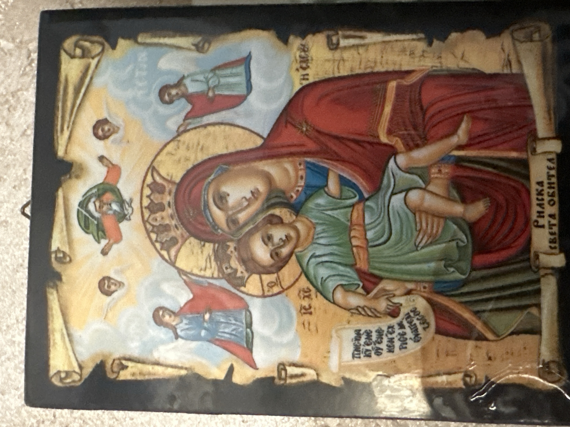 This is a photograph of an icon depicting the Virgin Mary holding the Christ child. They are surrounded by celestial figures and scrolls with writing. The icon is framed by a black border and has a dark, textured background. The style is traditional Orthodox Christian iconography. The photograph was taken in Marten, Bulgaria.