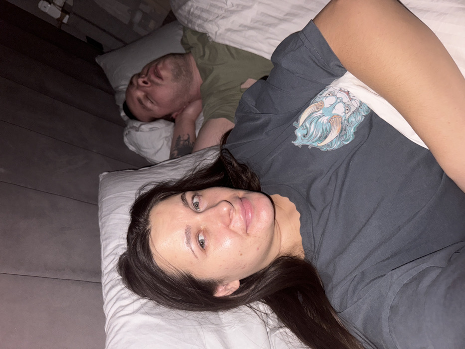A woman with long dark hair and light eyes lies on a white pillow, looking toward the camera. Her face is clear and slightly flushed. Behind her, partially under the covers, is a man sleeping on a separate pillow. He has dark hair, a short beard, and a tattoo is visible on his arm. The woman is wearing a dark gray t-shirt with a faded graphic design. The room appears dimly lit, with wooden flooring visible in the background.