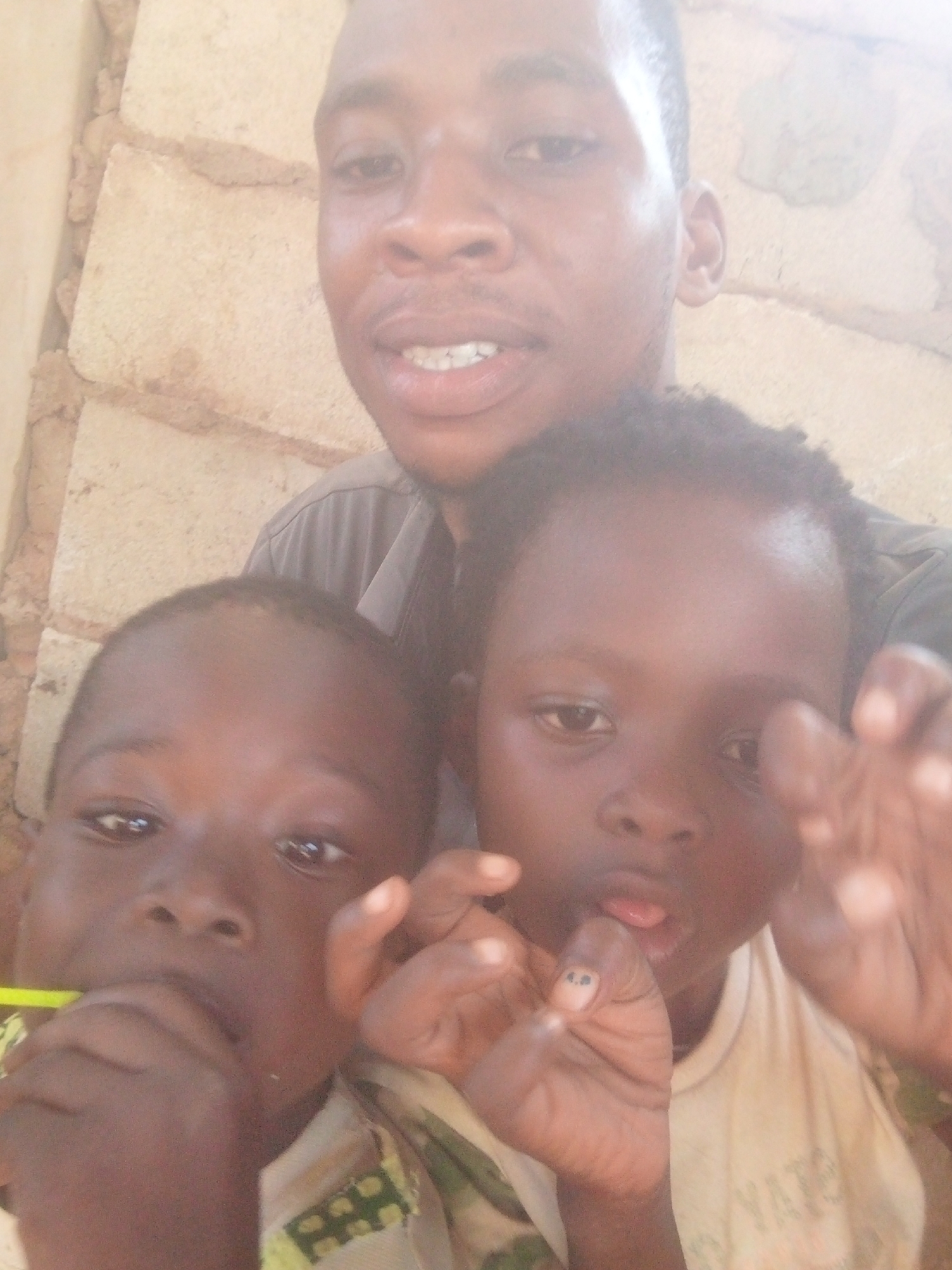 The image shows a man with two children. The man is standing behind the children, looking at the camera. The child on the left is holding a green object in his mouth. The child on the right is holding up his hand. The background appears to be a wall of stacked concrete blocks. The location is unknown.