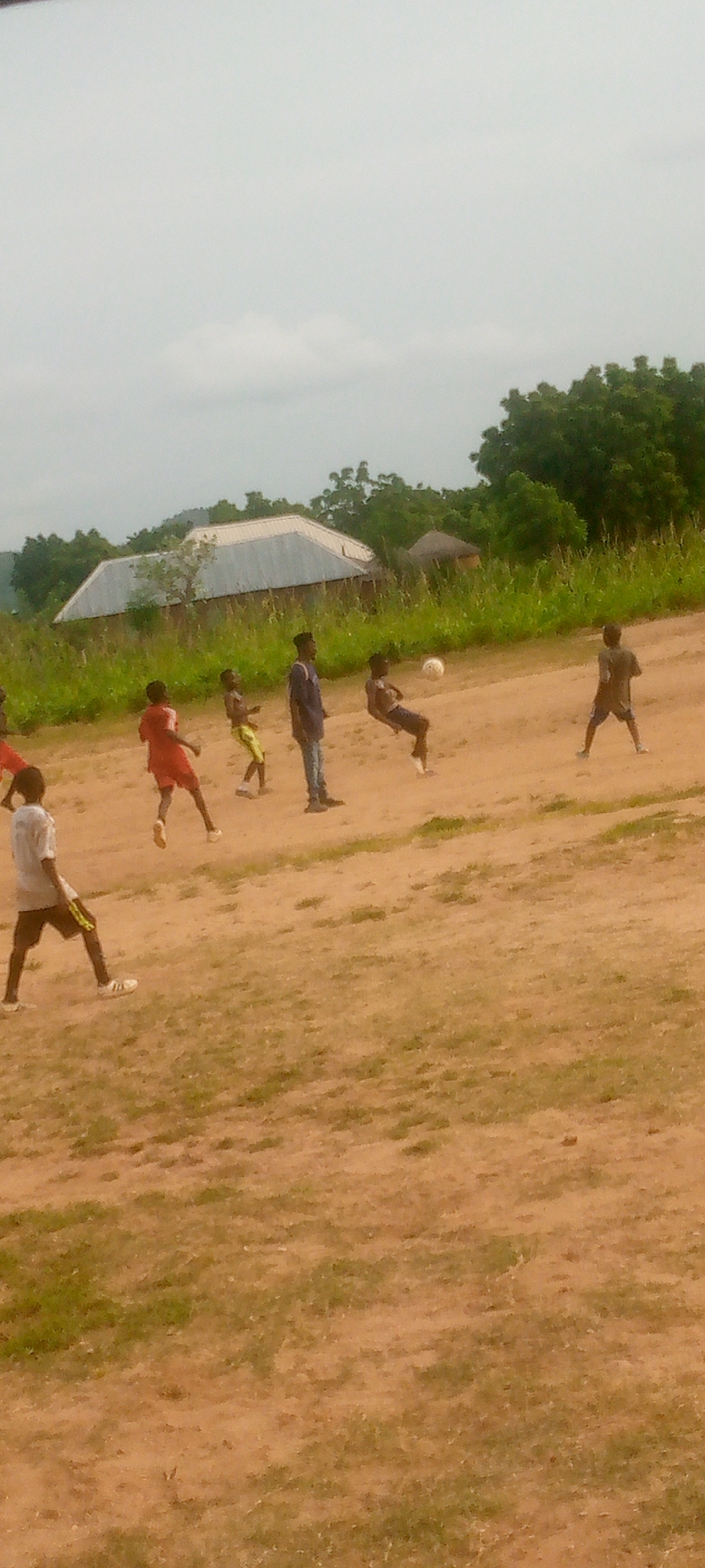The image depicts a group of children playing soccer on a dirt field in Langtang, Nigeria. The sky is overcast, and the ground is a mix of dry soil and sparse green grass. In the background, there are trees, some vegetation, and a few structures, including a building with a silver, corrugated metal roof and a smaller, round thatched hut.
There are at least six children visible. One child in a red shirt and shorts is in motion, appearing to kick a ball. Another child in a blue shirt and jeans is standing, watching the game. A third child, in dark shorts and a t-shirt, is captured mid-air, seemingly attempting a bicycle kick. A fourth child, wearing a light-colored shirt and dark shorts with yellow trim, is standing with one foot forward. Two other children are partially visible. The focus is on the soccer game and the surrounding rural environment.