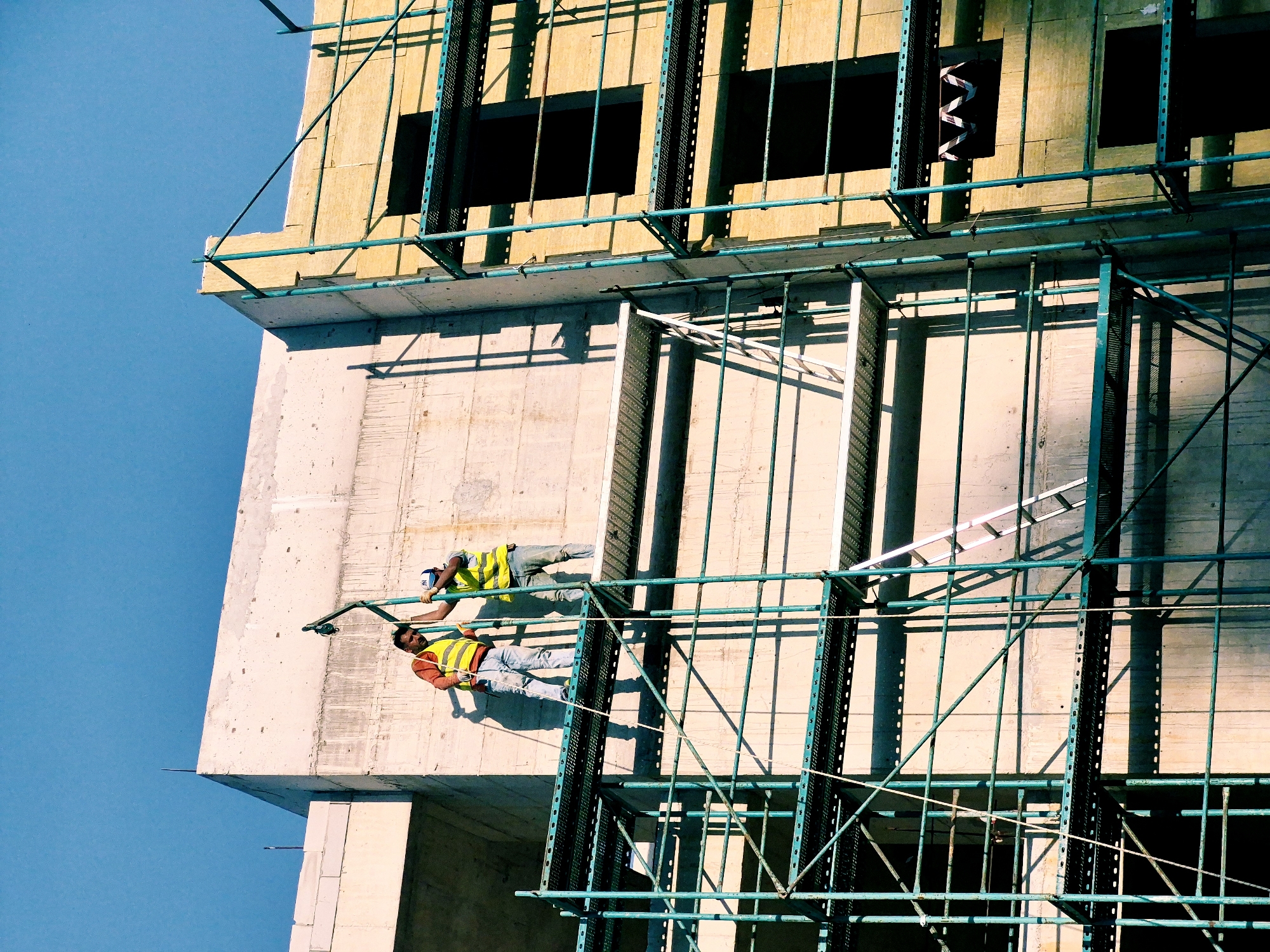 Two construction workers in high-visibility vests are working on the exterior of a building under construction. The building appears to be made of concrete and scaffolding, with wooden panels and metal supports. The scene is set against a clear blue sky, suggesting it is daytime. The workers are positioned at different heights, one above the other, and are secured by safety harnesses. They are using the scaffolding to access different parts of the building. One worker is leaning on a railing, while the other is holding onto a support beam. The sun is casting strong shadows on the building, indicating it is likely a sunny day. There are no visible signs of text in the image. The location is Đenovići, Montenegro.
