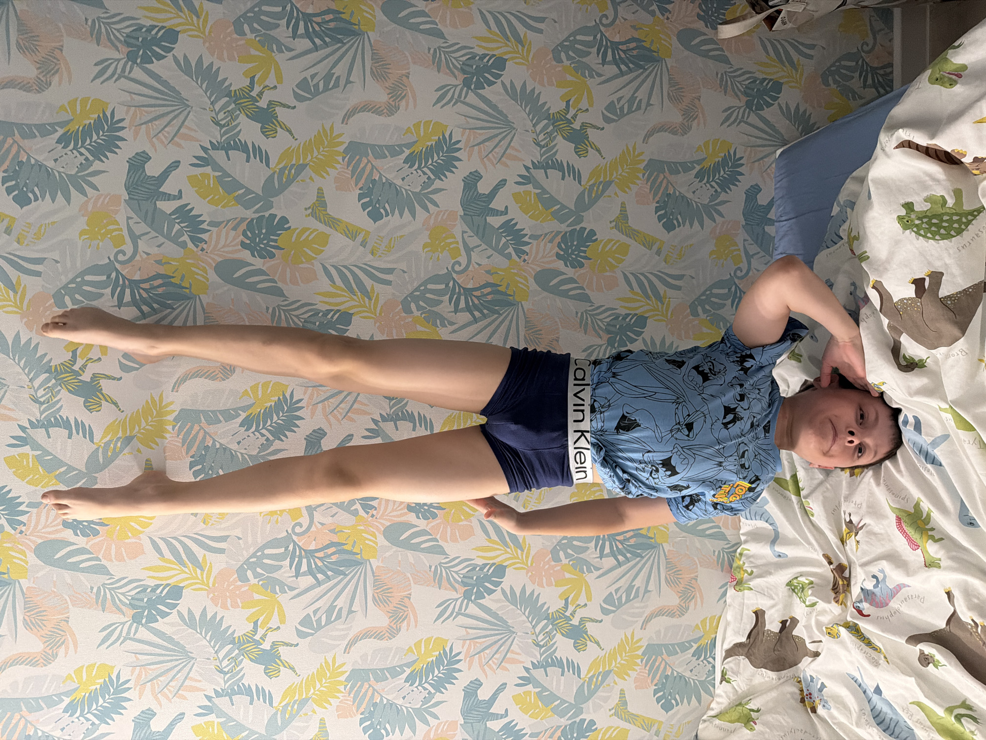 A person is lying on a bed. The individual wears blue boxer briefs with the "Calvin Klein" logo. They are also wearing a blue t-shirt with cartoon characters on it. Their legs are extended outward. The bed is covered with dinosaur-themed bedding. The wall is covered in wallpaper with a tropical print. The location is unspecified but the city is Votkinsk, Russia.