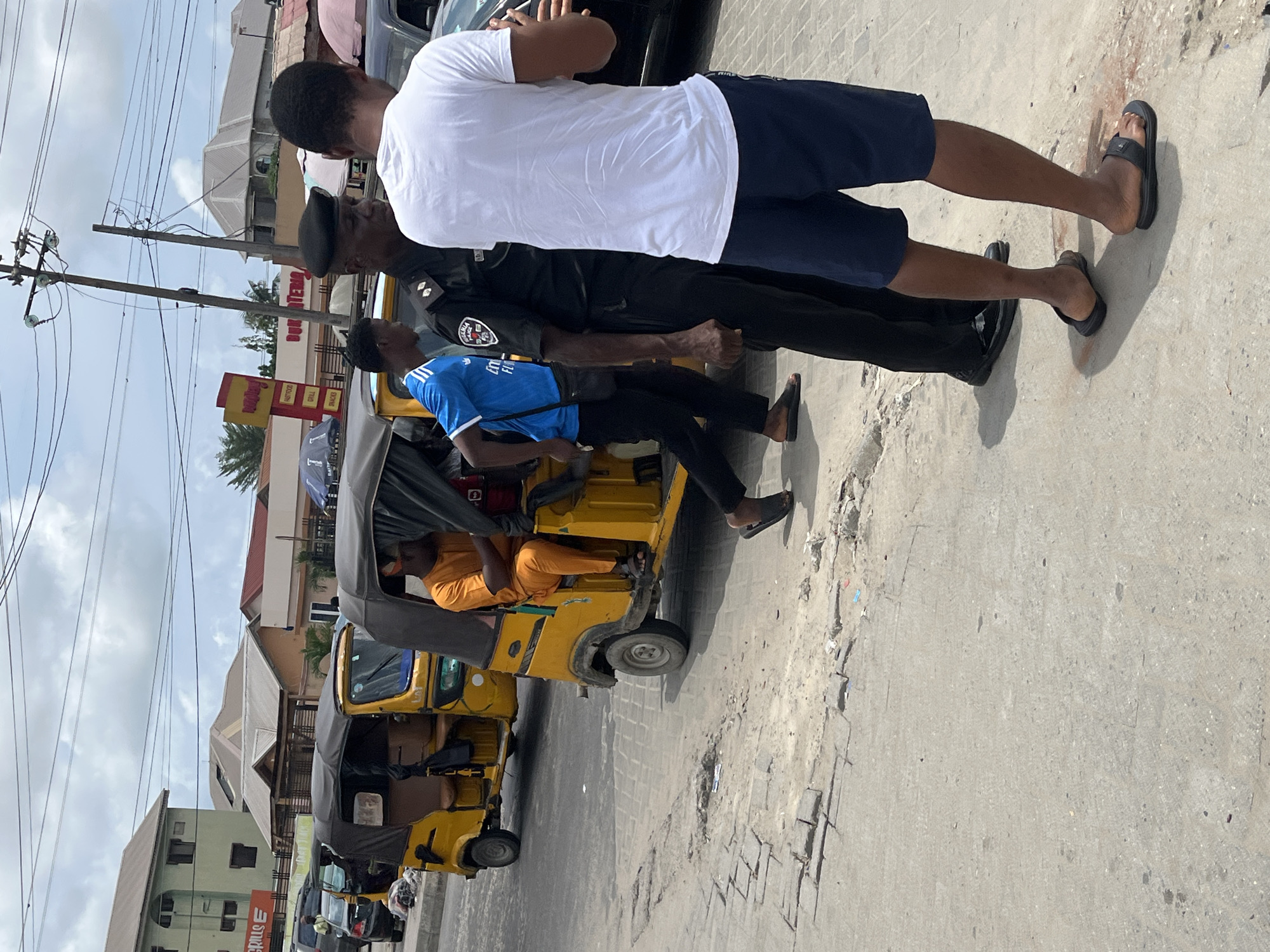 The image depicts a daytime street scene in Ado, Nigeria. In the foreground, a Nigerian Police Force officer in a dark green/black uniform and black beret stands, facing right, engaging with a civilian. This civilian, seen from behind, wears a white t-shirt, dark blue shorts, and black flip-flops, holding a white object in his left hand with his right hand also raised. To their left, another civilian, dressed in a blue t-shirt, dark trousers, and dark flip-flops, walks left, carrying a black shoulder bag.
On the road behind these individuals, two yellow Keke Napep tricycles with black canopies are visible; one has a passenger in an orange outfit seated inside. The concrete road surface and adjacent shoulder show signs of wear, cracks, and some scattered debris. In the background, various buildings line the street, including a multi-story light green structure and other single-story buildings with grey or brown roofs. A prominent red and yellow commercial sign advertises "Fastfood BUKATERIA GRILLS E", and another sign partially reads "DON'T TEM". Overhead, numerous utility lines connect to several wooden utility poles, set against an overcast sky. Other vehicles are partially visible on the road in the distance.