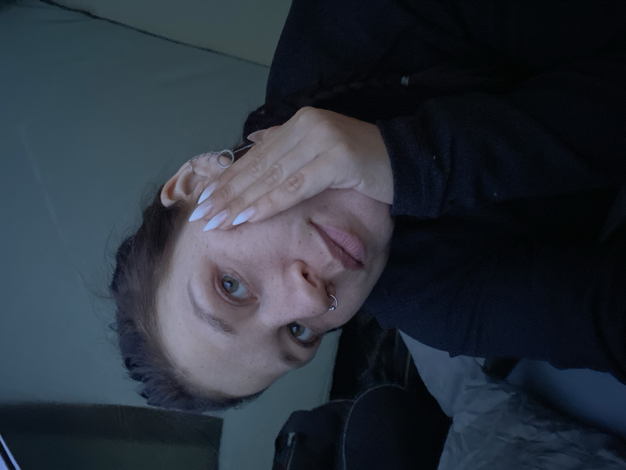 The image shows a close-up, self-taken photograph of a young woman's face, partially obscured by her hand. She has light skin with freckles, green eyes, and light pink lips. Her fingernails are long and painted white. She is wearing a dark-colored top and has a nose ring. In the background, a tent's interior is visible, with a sleeping bag or mat on the left. The overall lighting is dim, suggesting an indoor or low-light environment. The location is identified as Абзаново, Russia.