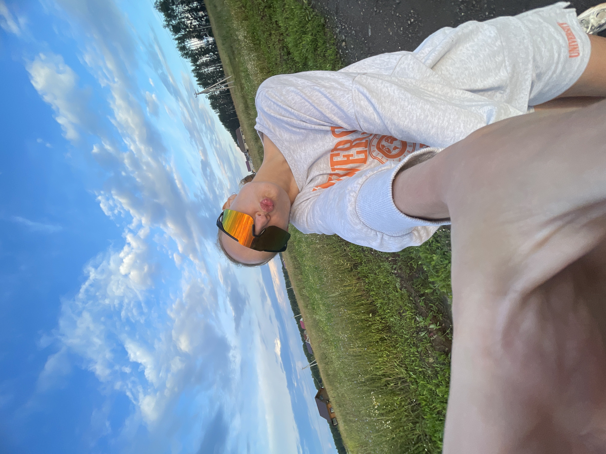 The image is a selfie taken outdoors in a rural area, likely in or near the village of Podshivalovo, Russia, given the context. The background features a vast field of green grass and wildflowers under a blue sky with scattered white clouds. In the distance, several residential houses with red roofs are visible amongst scattered trees. A line of coniferous trees forms a dense forest on the horizon. There are also utility poles and wires in the distance. The ground in the foreground is a dirt path.
In the foreground, a person is captured from the chest up, holding the camera with their left hand, which is prominently in the bottom left of the frame. The person is wearing a light grey sweatshirt with orange lettering and graphics, and matching light grey shorts. They are also wearing oversized sunglasses with a reflective orange and yellow tint. The person's head is tilted, and their lips are pursed in a kissing gesture, directed towards the camera. The lighting suggests it is either early morning or late afternoon, with the sun casting a warm glow.