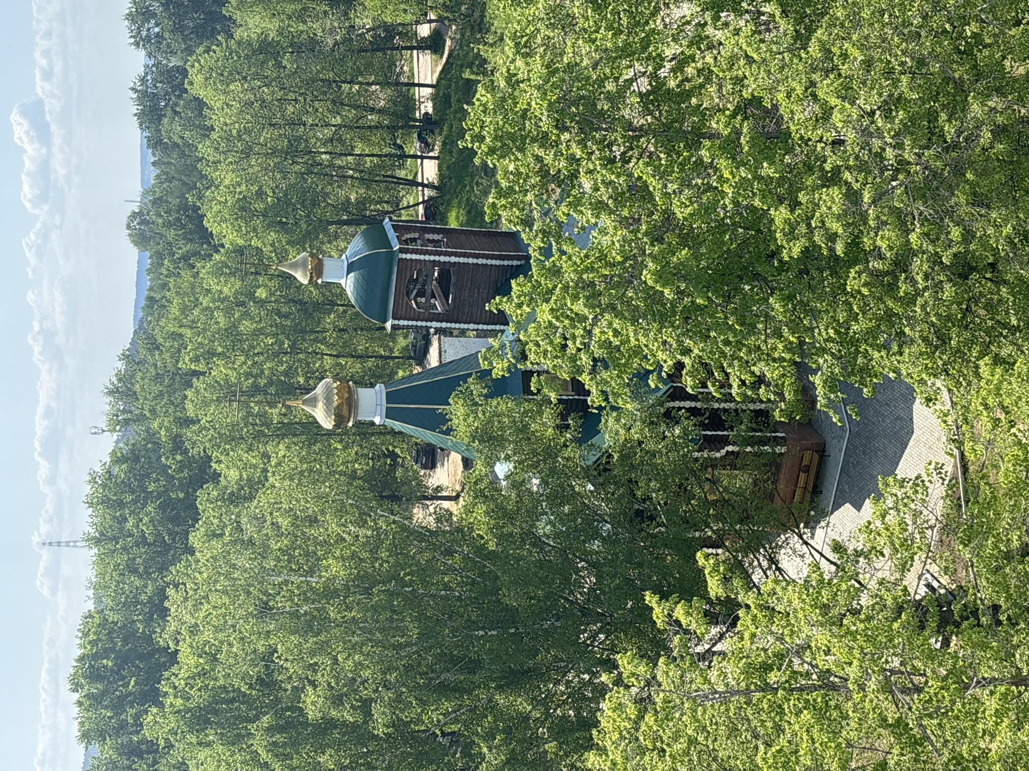 The image shows a view of the Church of St. Nicholas, located in the city of Chaikovsky, Russia. The church has two domes, one larger than the other, and a wooden bell tower. It is surrounded by trees with lush green foliage, partially obscuring the structure. A paved pathway and a bench are visible next to the church. The sky in the background is pale blue with some clouds. A tall transmission tower is also seen. The image is taken from a high vantage point.