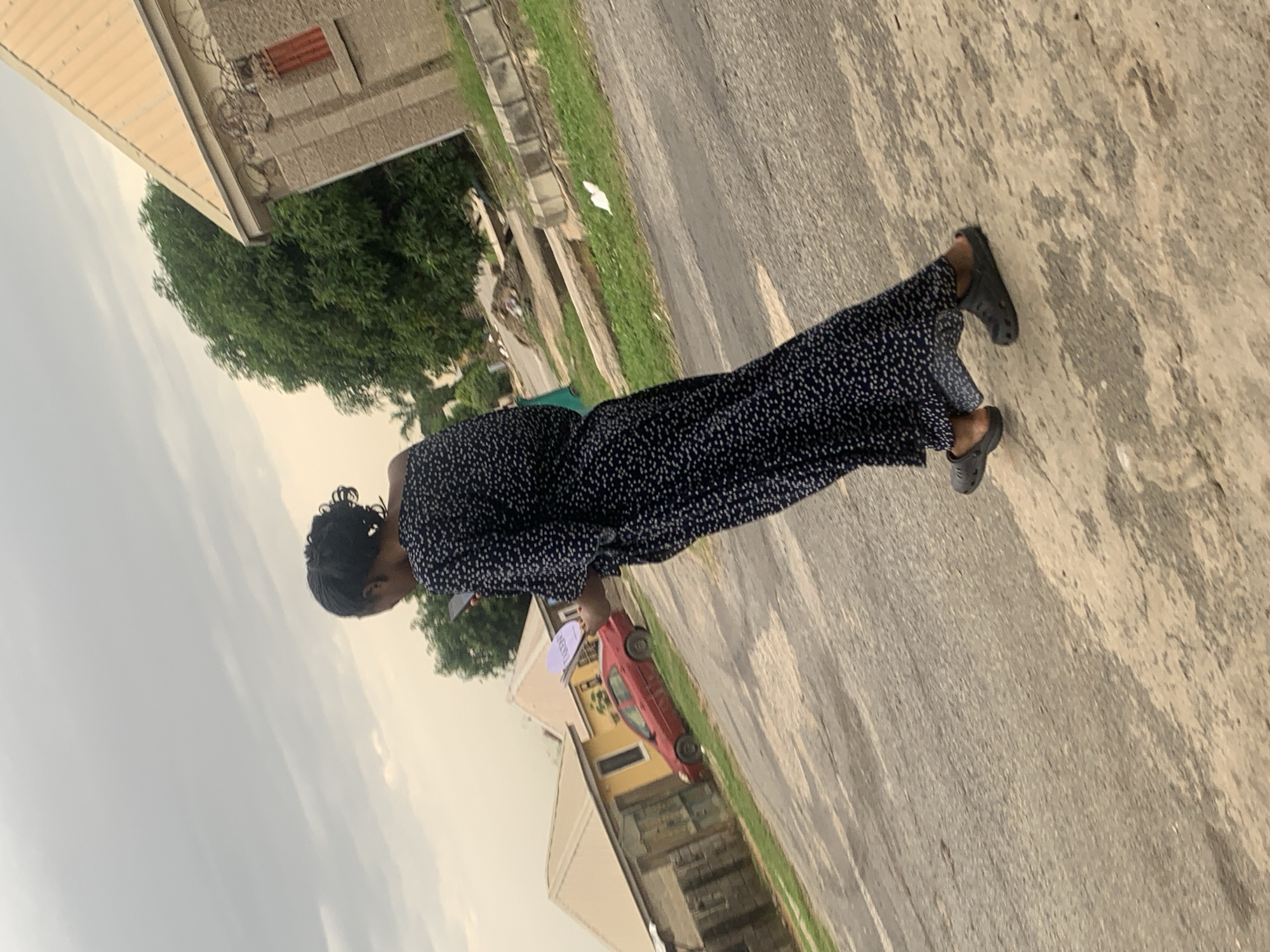 A woman, seen from behind and in profile, walks away from the camera on a paved street, holding a light purple object in her right hand. She is wearing a long, dark blue dress with a white polka dot pattern and dark gray clogs. The street is lined with grass and concrete drainage ditches. In the background, there are two buildings with yellow and cream-colored walls and beige roofs, and a red car parked on the side of the road. A large green tree stands between the buildings. The sky is overcast with pale clouds. The scene appears to be in a residential area, possibly in the city of Abuja, Nigeria.