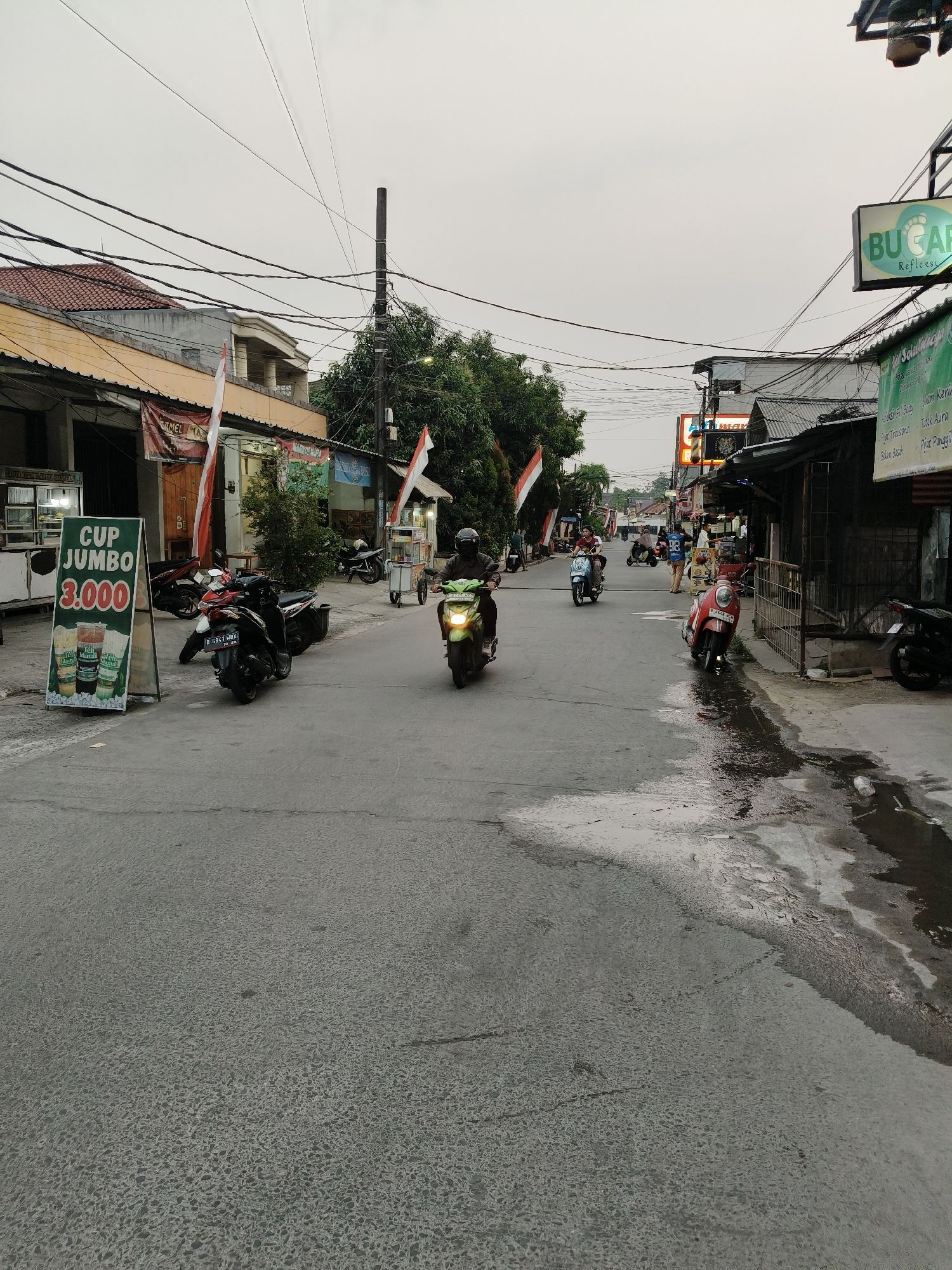 The image depicts a street scene in the city of South Tangerang, Indonesia, likely during overcast weather as indicated by the muted sky. The street is paved and shows signs of recent wetness, with several puddles reflecting the surrounding environment. A variety of buildings, commercial establishments, and residential structures line both sides of the street, exhibiting a mix of architectural styles and construction materials.
Several individuals are present, primarily riding motorcycles and scooters, suggesting it's a common mode of transportation in this urban setting. One person on a green scooter is prominently featured in the foreground, heading towards the viewer. Other motorcycles are parked along the roadside, and a few more are seen further down the street, indicating local commerce and daily activities.
Notable objects in the scene include signage for various businesses. One prominent sign displays "CUP JUMBO 3.000" along with images of beverages, indicating a food or drink vendor. Another sign, partially visible, reads "BUCAR Refleksi," suggesting a reflexology or spa service. Street lights and overhead electrical wires are abundant, characteristic of urban infrastructure. Indonesian flags are displayed on poles, indicating a national presence.
The overall impression is one of a busy, everyday street in a developing urban area of Indonesia, with a focus on small businesses and local transportation.