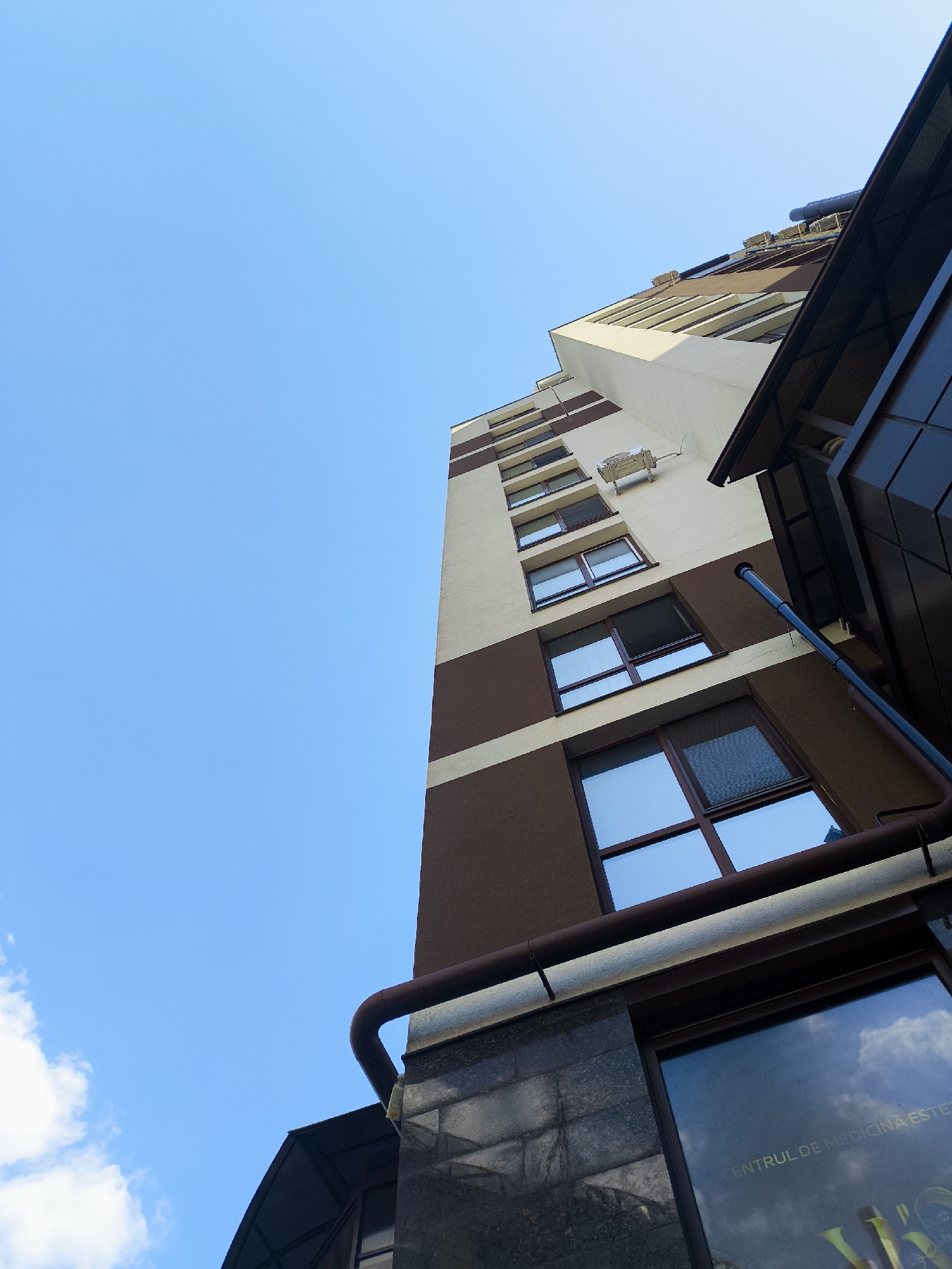 The image shows a low-angle view of a modern apartment building against a clear blue sky. The building has a contrasting facade of light cream and dark brown sections, with rows of windows. A dark brown gutter system runs along the side of the building. In the foreground, a darker, reflective surface, possibly part of a ground-level shop or entrance, shows reflections of the sky and clouds, and text that reads "CENTRUL DE MEDICINA ESTE". There are no people or animals visible in the image. The environment appears to be an urban setting on a clear day.
