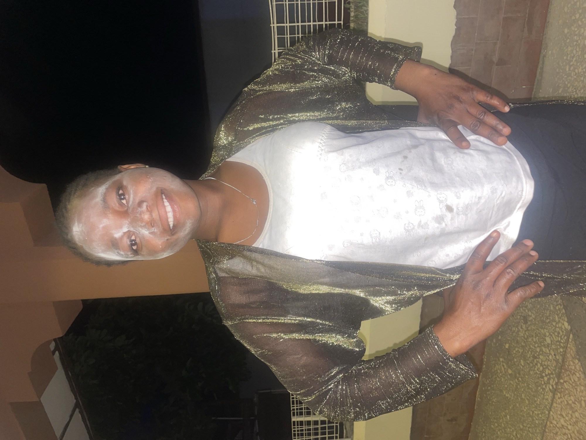 A medium indoor shot shows a woman with a thin necklace, white shirt, black bottoms, and a gold, sparkly mesh jacket, facing the camera with her hands on her torso. She has a white cream-like substance on her face, from her hairline to just under her eyes and cheekbones. She is smiling. The environment is a dimly-lit area, possibly a building exterior in Jos, Nigeria. Behind her is a combination of dark space, a lit beige structure, and greenery. A barred window can be seen on the upper right. The surface on which she stands is a beige mottled material, possibly tiled.