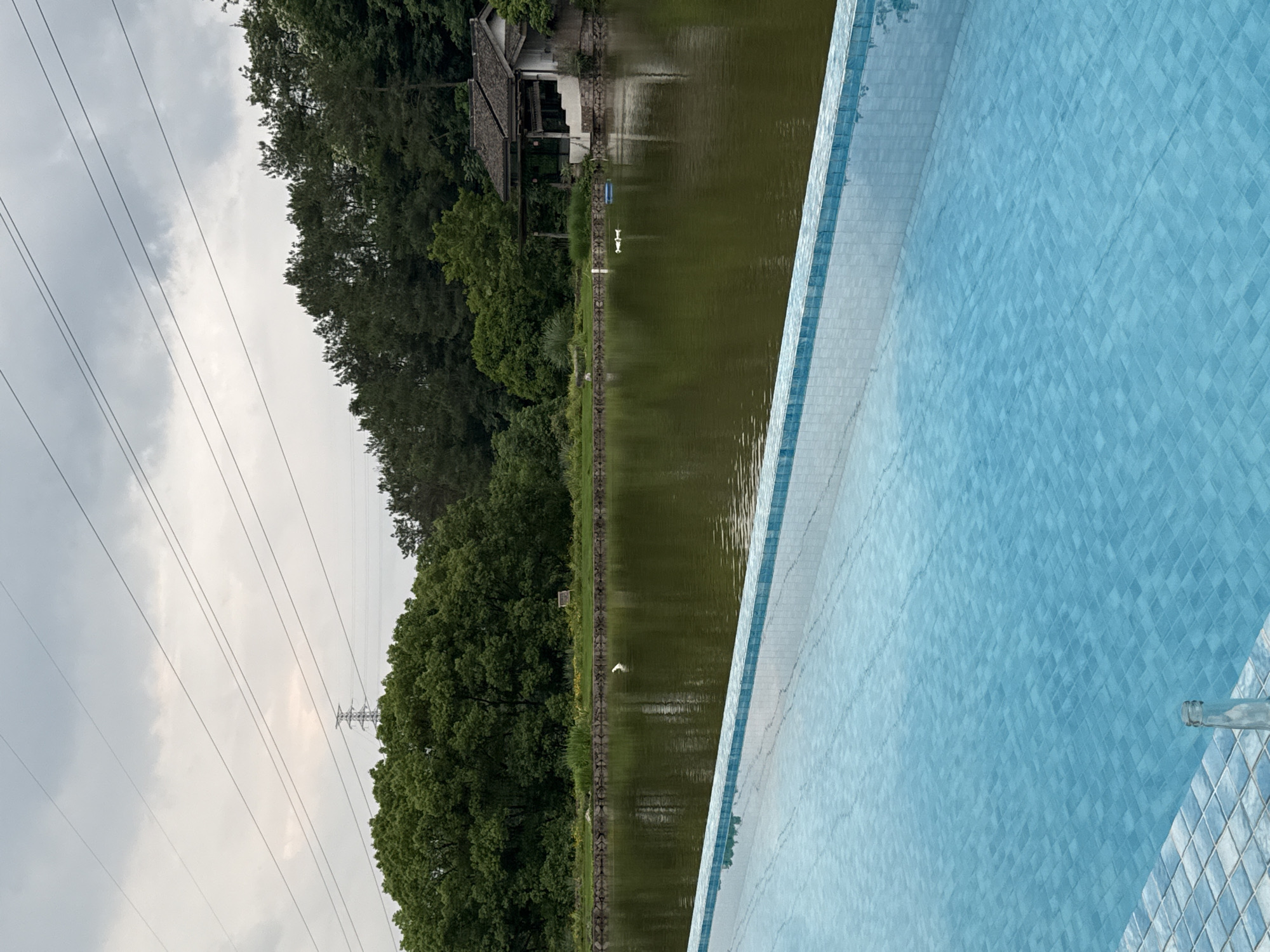 The image presents a view of a serene landscape, likely in a rural area. A body of water, possibly a lake or large pond, stretches across the midground, its surface reflecting the surrounding environment. Along the water's edge, a fence runs parallel, separating the water from a dense line of trees and greenery. A small structure, resembling a building or hut, is visible on the opposite bank of the water.
In the foreground, there's a section of a blue-tiled wall, suggesting a pool or other man-made structure. Overhead, electrical power lines stretch across the sky, supported by a utility pole. The sky is overcast with cloudy skies. There is no indication that the photograph was taken in Jinhua, China.
