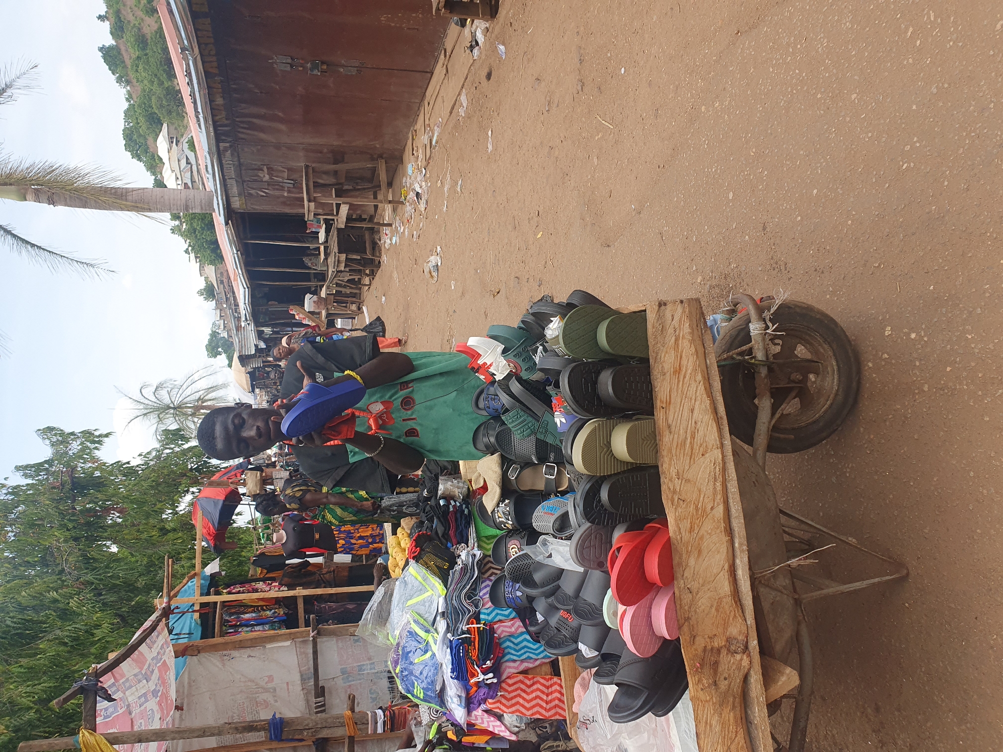 A street vendor stands next to a wheelbarrow filled with sandals for sale, in Lokoja, Nigeria. The vendor is holding up a blue sandal. His wheelbarrow is filled with various colorful sandals. The background includes other market stalls and a dirt road.