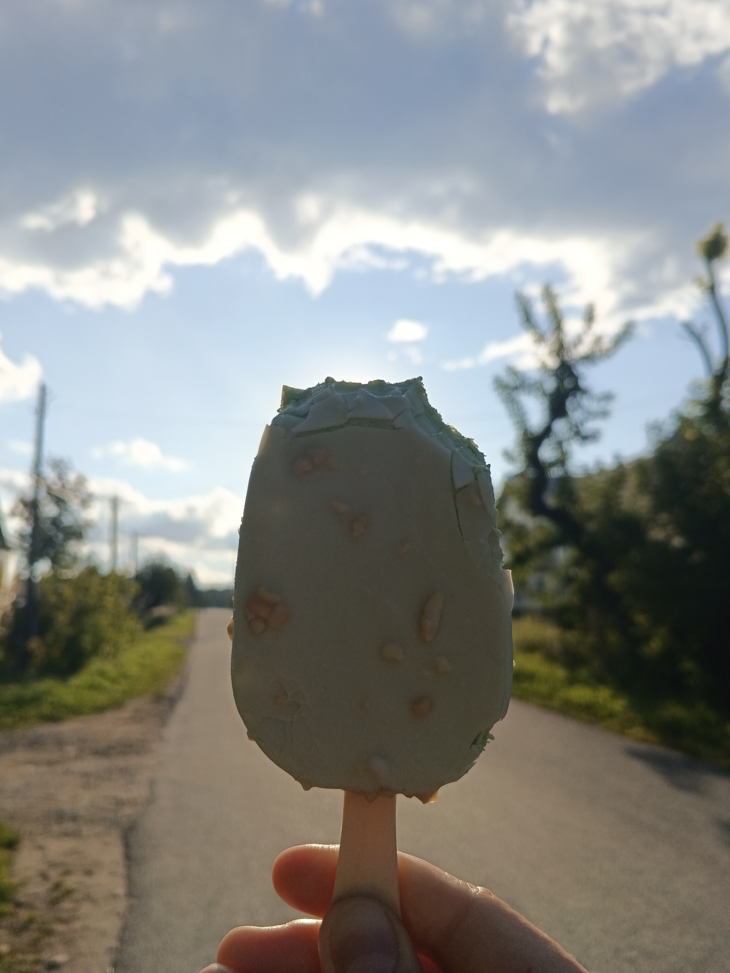 A close-up, low-angle shot captures a hand holding a partially eaten, light green ice cream bar with visible nut pieces. The ice cream bar is positioned centrally in the frame, with its stick extending downwards. The background features a rural road lined with green vegetation and trees, leading towards a hazy horizon under a cloudy sky. The overall setting suggests a warm, outdoor environment. The location is identified as Medvedyovka, Russia.