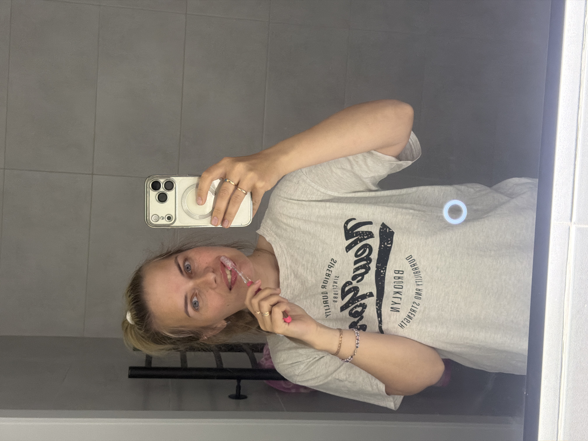 The image depicts a fair-skinned woman, appearing to be in her 20s or 30s, reflected in a large mirror within a bathroom. Her blonde hair is tied back in a high ponytail with a white scrunchie. She has blue eyes and a slight smile, with light freckles visible on her nose and cheeks.
She is wearing a light grey short-sleeved t-shirt with a mirrored black graphic print. When reversed, the text on the t-shirt reads "SUPERIOR QUALITY," "EST. 1989," "New York" in a large script font, and "BROOKLYN," "DURABILITY AND STRENGTH."
The woman is actively brushing her teeth with a pink toothbrush, which is partially inserted into her mouth, showing some white toothpaste foam. She holds a light-colored smartphone, identifiable as an iPhone due to its triple-lens camera array and visible MagSafe ring on the back, in her right hand to capture the mirror selfie.
On her left wrist, she wears two bracelets: one thin, gold-colored chain and one beaded bracelet with purple/blue and silver elements. Several gold-toned rings are visible on the fingers of her left hand.
The background, as reflected in the mirror, consists of a grey tiled wall with horizontal rectangular tiles and visible grout lines. To the left of the woman's reflection, a dark-colored towel warmer or drying rack is visible, with what appears to be a light-colored (possibly pink) garment hanging from it. A subtle horizontal light bar runs along the bottom edge of the mirror, and a faint, glowing white-blue circular light is visible on the mirror's surface in the lower right quadrant.
The image itself contains no discernible visual markers such as signage, unique architectural features, or geographical identifiers to confirm the specified location of Chaikovsky, Russia. The reflected t-shirt graphics reference "New York" and "Brooklyn."