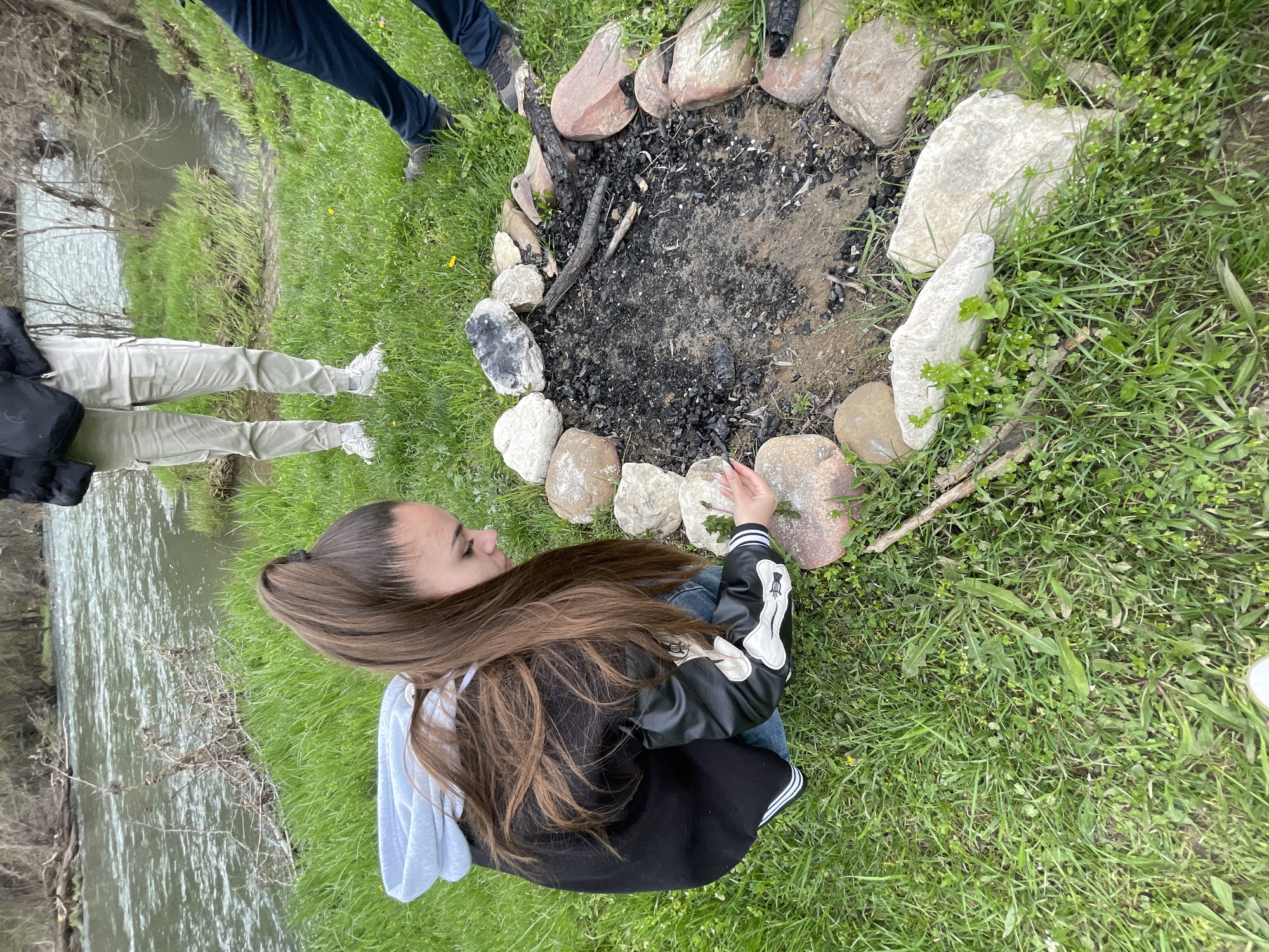 A young woman with long brown hair is kneeling on green grass next to a ring of stones surrounding a fire pit filled with ashes and charred wood. A river or stream is visible in the background to the left. Two other individuals are partially visible, one with dark blue pants and shoes, and another with beige pants and white shoes, standing near the fire pit. The location appears to be outdoors, near a body of water, in a grassy area. The photo does not contain sufficient information to determine if it is in Barkach, Bulgaria.