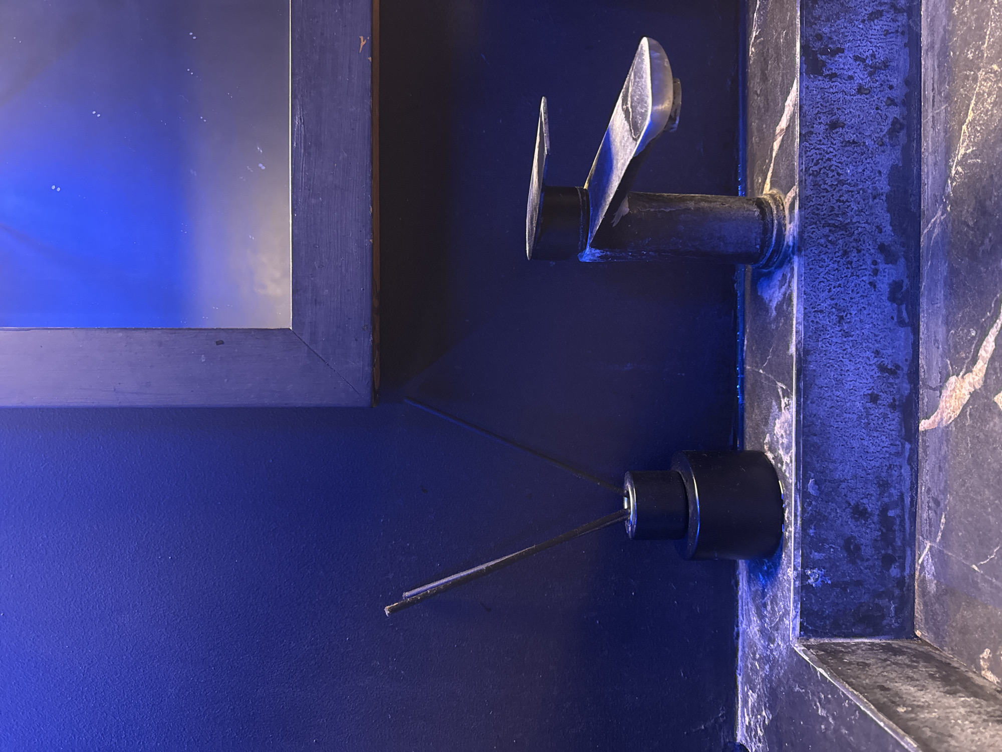 The image displays a close-up view of a bathroom sink area, possibly in Kuta Selatan, Indonesia, given the context. A dark, textured wall serves as the backdrop. To the left, a black reed diffuser with two dark reeds stands on a dark marble countertop. In the center-right, a metallic faucet with a modern design is positioned on the same countertop. The faucet appears somewhat aged or stained. Above the countertop and faucet, a dark-framed mirror reflects a blue, possibly illuminated, surface and some indistinct shapes. The overall lighting is dim and casts a blue hue over the scene, suggesting artificial lighting or a specific color ambiance. The marble countertop shows variations in its pattern and texture, with some lighter veining visible. The scene is static, with no people or apparent activity.