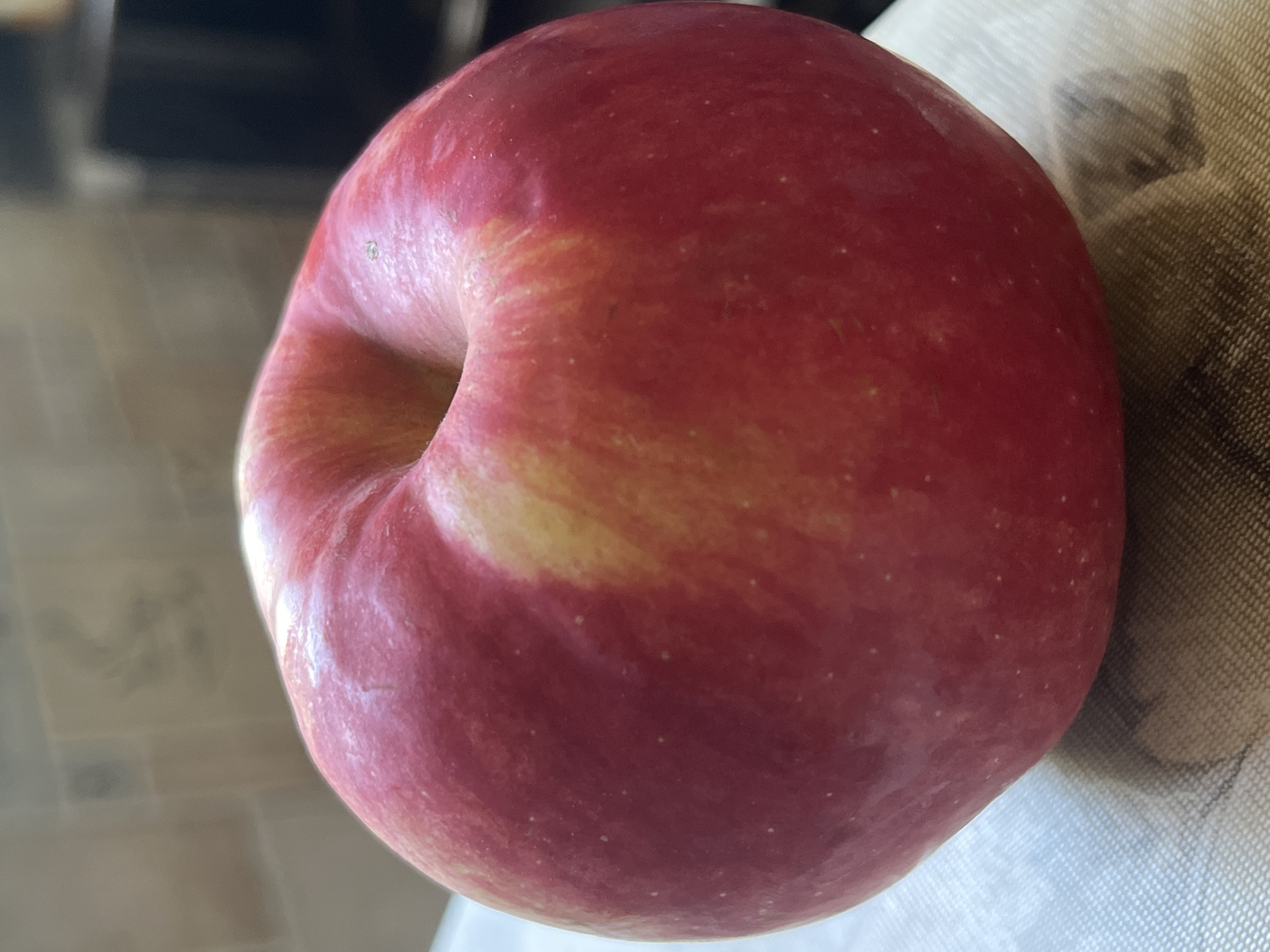 The image shows a close-up of a single red apple. The apple is round with a slight indent at the top where a stem would be. It has streaks of yellow and lighter red mixed with its dominant deep red color. The apple is resting on a textured surface, possibly a placemat or a cutting board, which has a light-colored, patterned texture. In the blurred background, the environment appears to be indoors, with hints of a kitchen or dining area. There are no people or specific landmarks visible that would indicate a location in Minsk, Belarus.