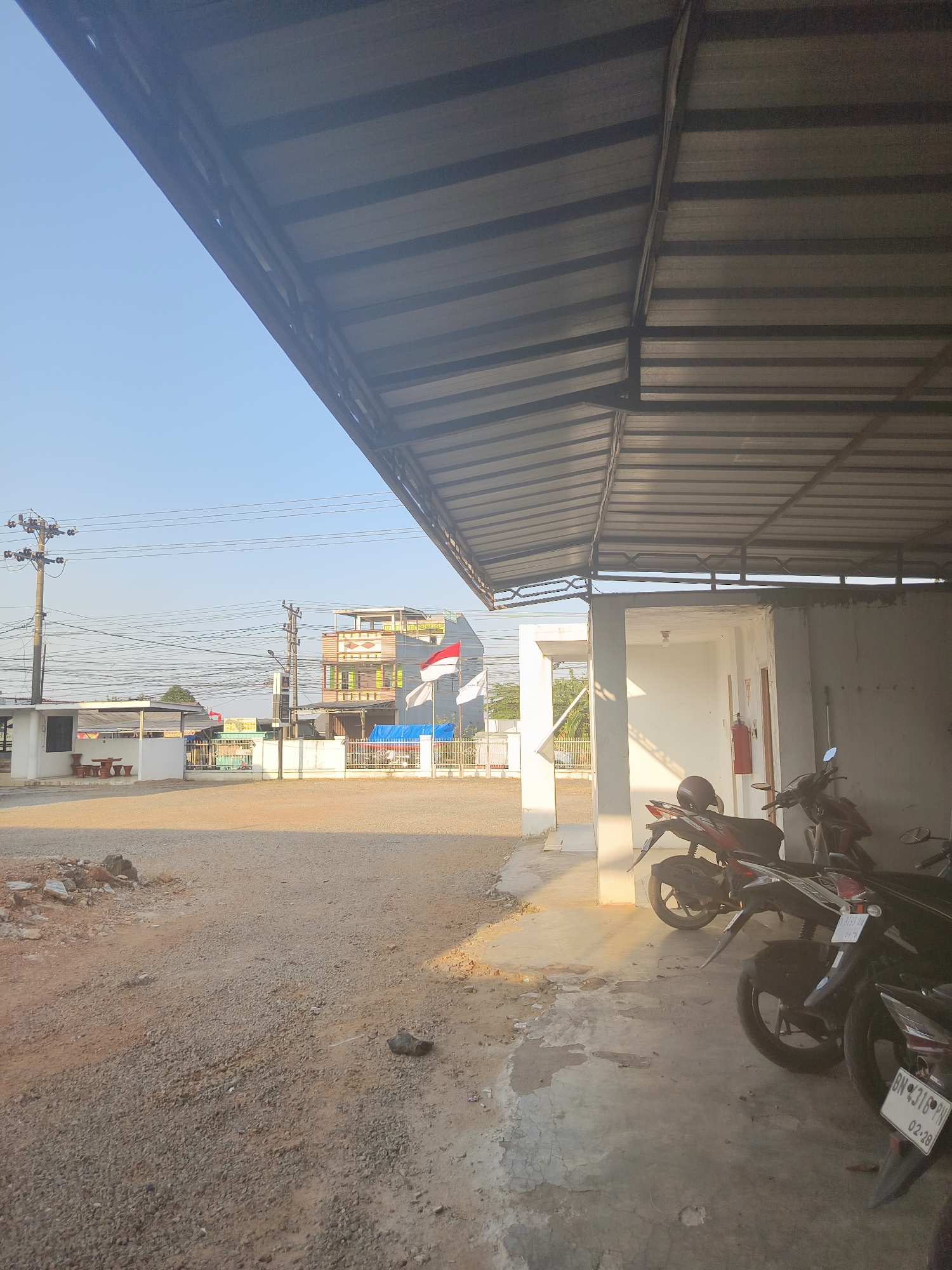The image depicts an outdoor scene in Pangkalpinang, Indonesia, under a clear blue sky. A corrugated metal roof structure extends over a concrete area where several motorcycles are parked. To the left, a gravel-covered lot transitions to a paved area in front of white buildings and a small outdoor seating arrangement. In the background, a multi-story building with colorful accents is visible, adorned with Indonesian flags. Power lines and utility poles are also present in the mid-ground, suggesting an urban or semi-urban environment. The lighting indicates it is either early morning or late afternoon, with warm sunlight casting shadows. There are no people visible in the frame.