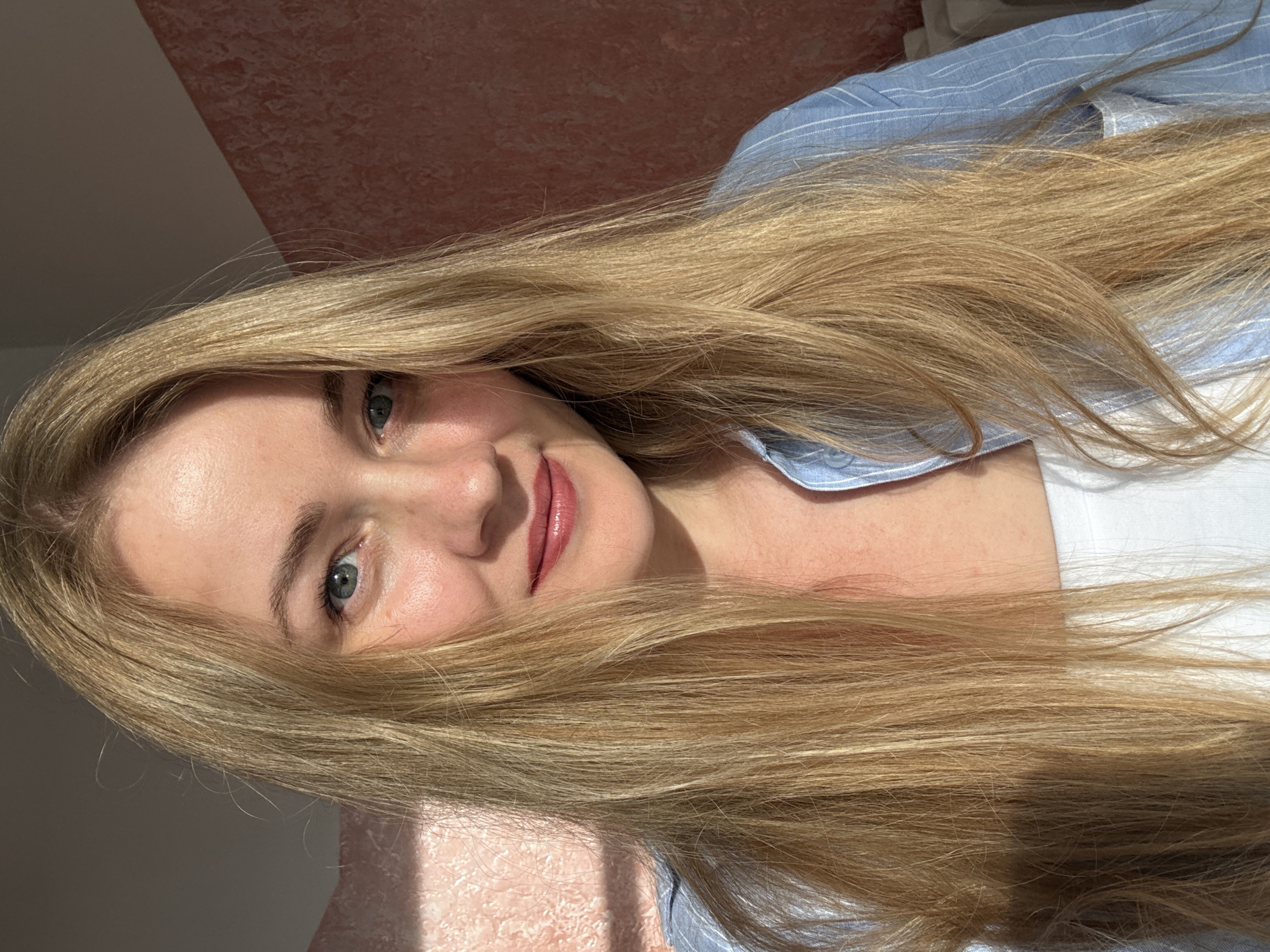 A woman with long blonde hair is shown in a close-up portrait. She is wearing a blue striped shirt and a white top underneath. The background appears to be an indoor setting with a pink textured wall and a hint of natural light casting shadows. The woman's face is directed towards the camera, with her eyes looking slightly to the left. Her lips are parted in a subtle smile, and she is wearing lip gloss. There is no specific indication of the city of Chaikovsky, Russia within the image itself.
