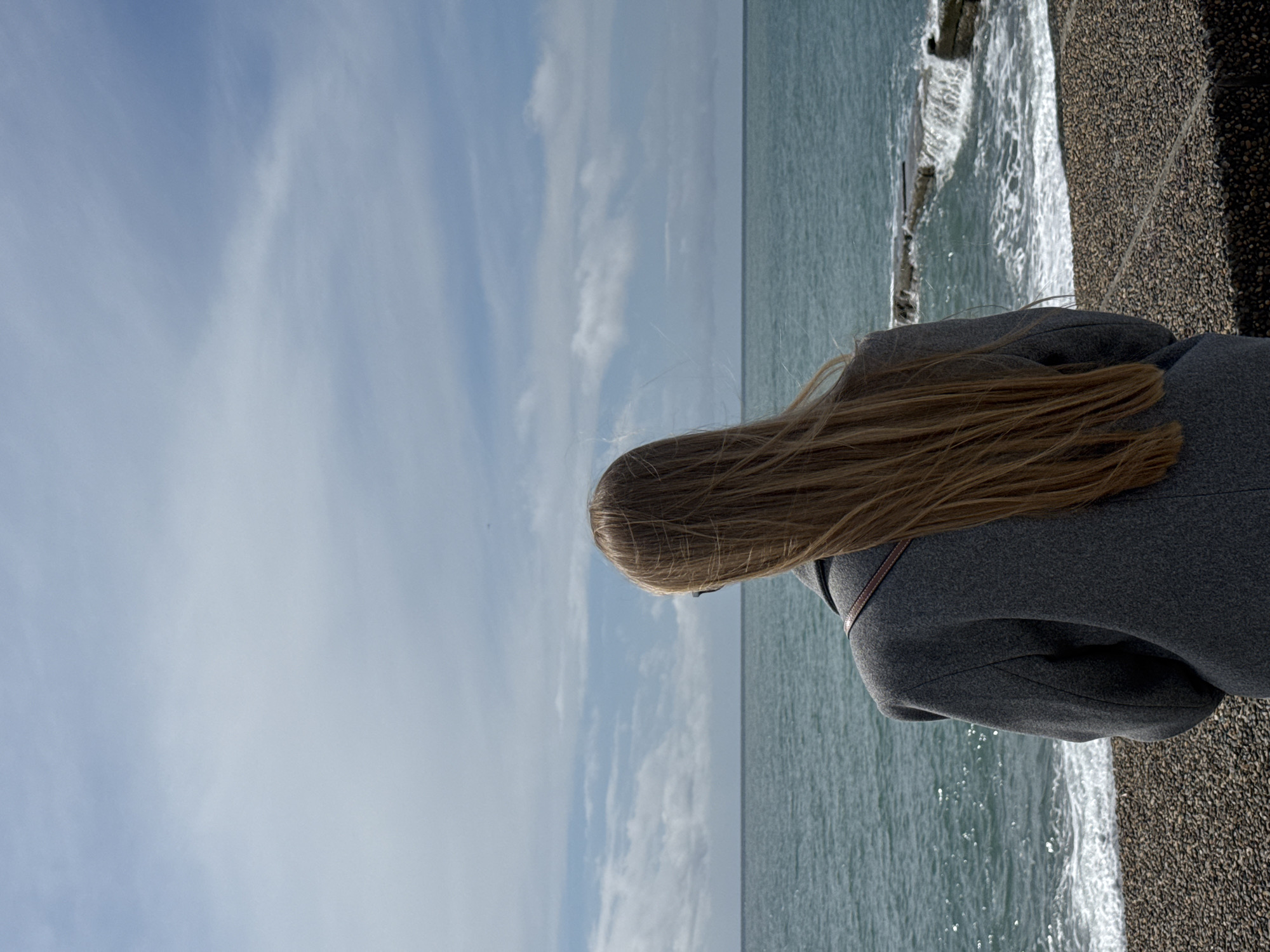 A woman with long blonde hair, wearing a gray coat, is standing near the seashore. The location is Sochi, Russia. The woman is looking at the sea, which is stretching out to the horizon under a blue sky with streaks of clouds. The waves are breaking on the shore, which is composed of small pebbles.