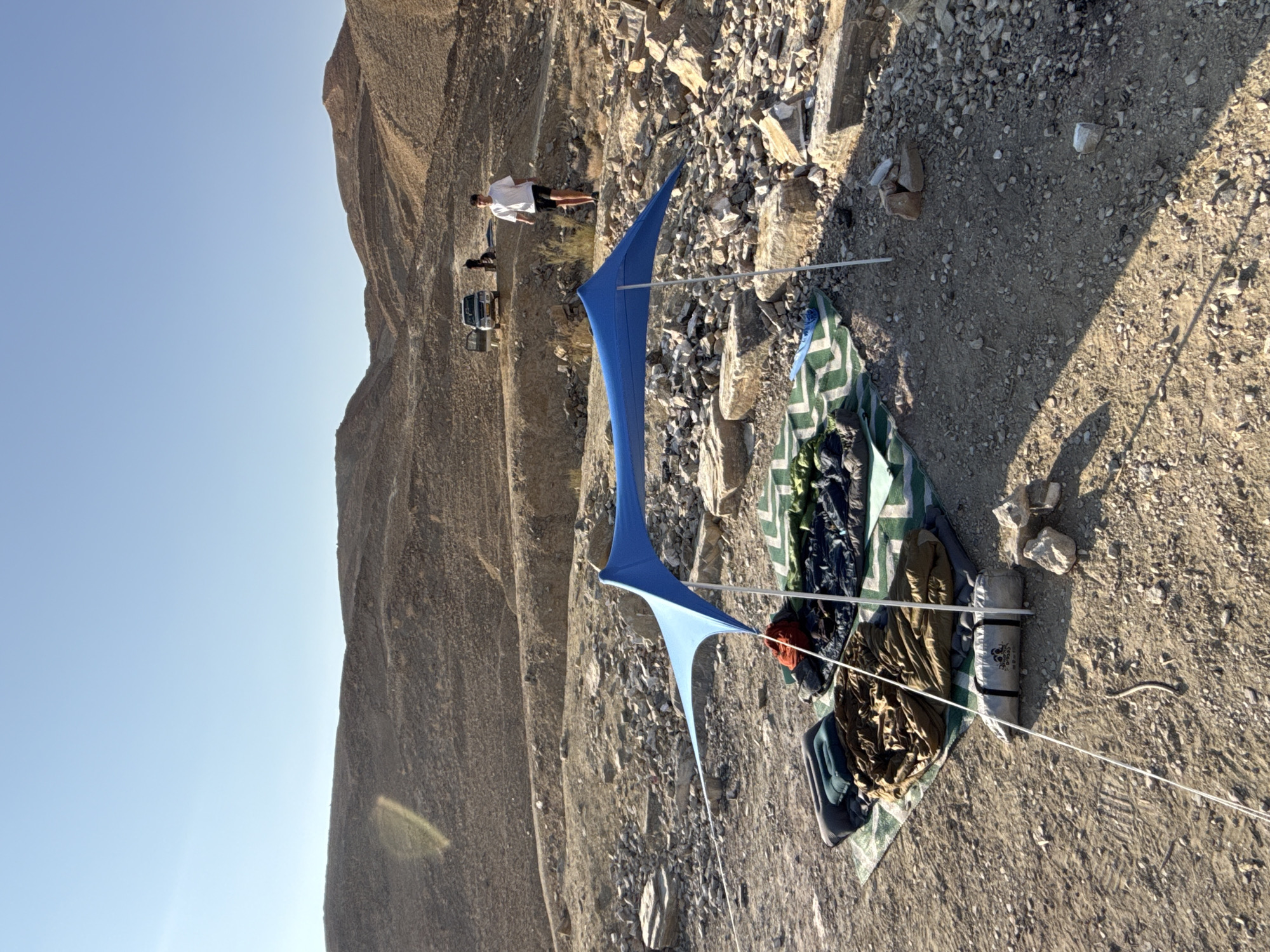 In the arid landscape of the Ramat Negev Regional Council, Israel, a campsite is set up in a rocky, barren terrain under a clear blue sky. A blue canopy tent is pitched over a green and white striped mat, upon which several sleeping bags and sleeping pads are laid out. A rolled-up sleeping bag is also visible in the foreground. In the middle ground, a person in a white t-shirt and dark shorts walks away from the camera towards a white SUV parked on a dirt track in front of a hilly, rocky backdrop. Another person is partially visible near the SUV. The environment is characterized by sparse vegetation, sand, and numerous rocks and boulders scattered across the ground, with rolling hills dominating the background.