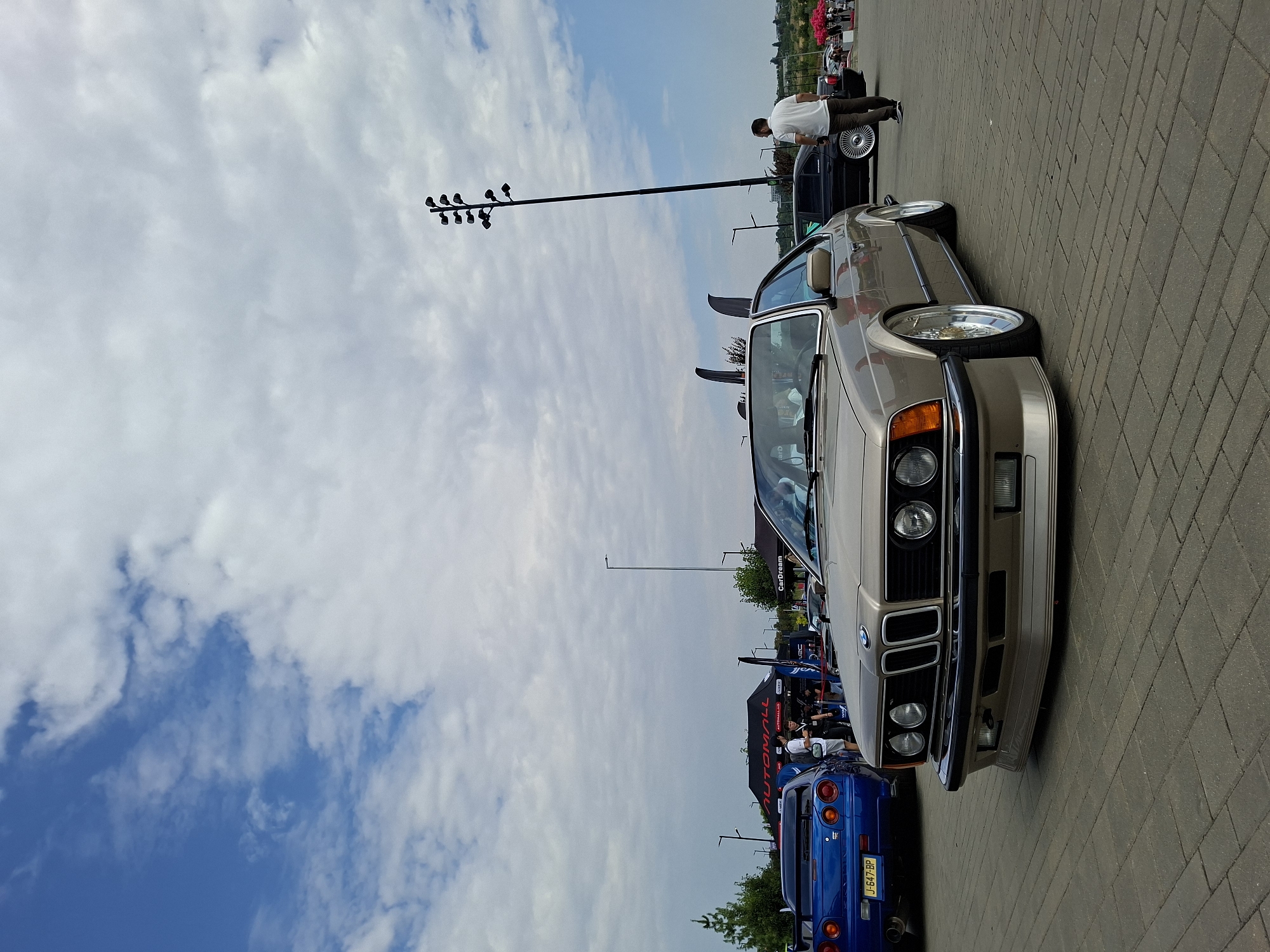 In the city of Stăuceni, Moldova, a beige BMW E24 coupe is prominently displayed on a paved lot under a cloudy blue sky. The car is lowered, with custom chrome rims. To the left, a blue Nissan Skyline R34 GT-T with a large rear spoiler is parked. Behind the BMW and Skyline, several tents are visible, with people milling around. A tall, multi-headed lamp post stands in the background. The scene appears to be an outdoor car show or event.