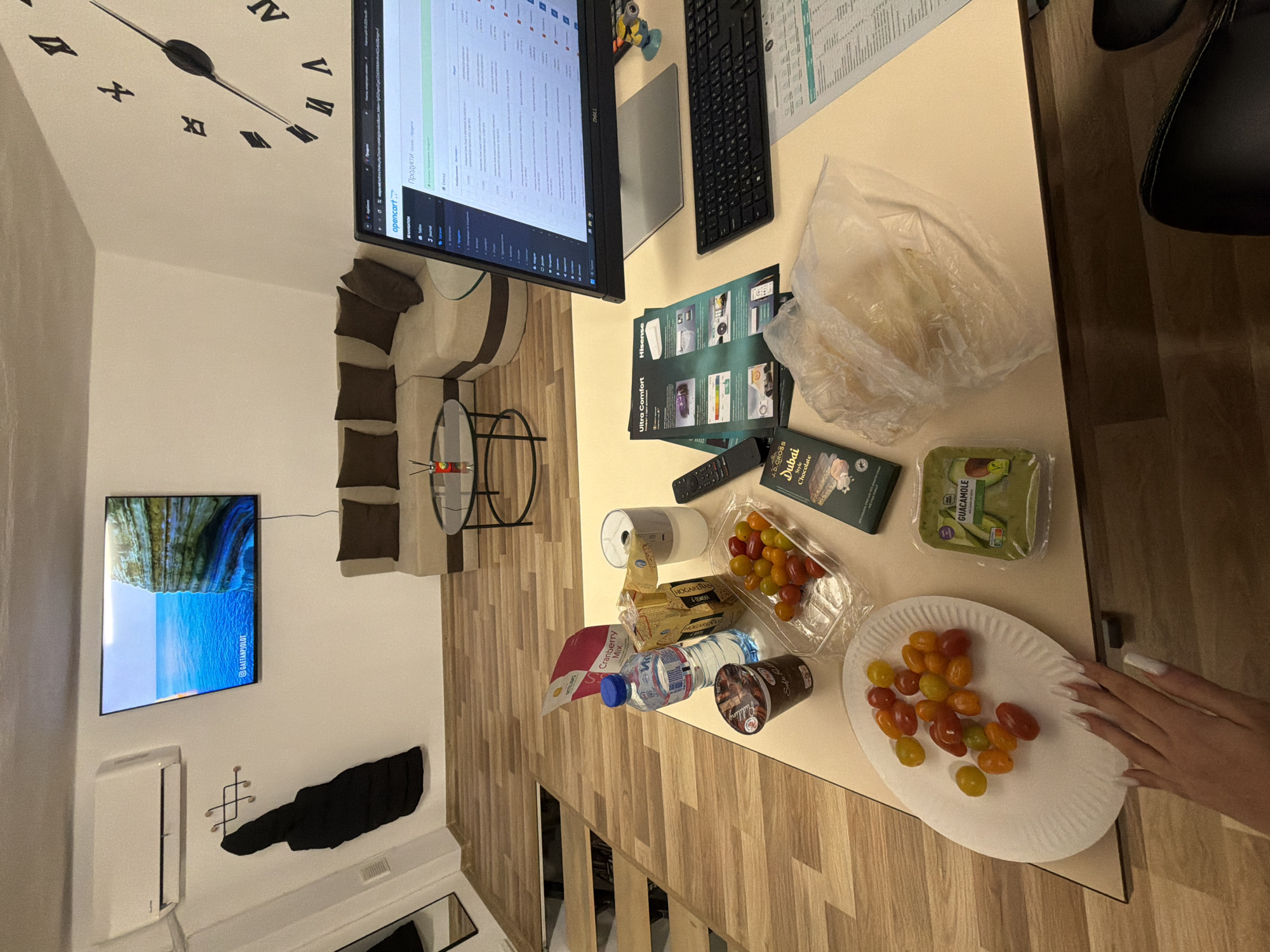 The image shows a cluttered desk in what appears to be an indoor setting, potentially a home office. A computer monitor is centrally positioned, displaying a page with text and data, possibly a website or application interface. Adjacent to the monitor is a keyboard and a tablet.
Scattered across the desk are various items, including a plastic bag filled with food, a container of cherry tomatoes, a package of guacamole, a box of chocolate, a remote control, a roll of paper towels, a package of crackers, a bottle of water, and a cup of yogurt. There's also a stack of papers with printed images and text.
A hand with long, white-painted nails is visible in the lower right corner, seemingly interacting with the plate of tomatoes.
The background reveals a living area with a wall-mounted television displaying a scenic landscape. There is also a clock on the wall, a couch, and a coffee table. The flooring appears to be wooden planks.
The location information provided (Gabrovo, Bulgaria) does not correlate with the visual data in the image, as it is not possible to determine the location based solely on the contents of the photograph.