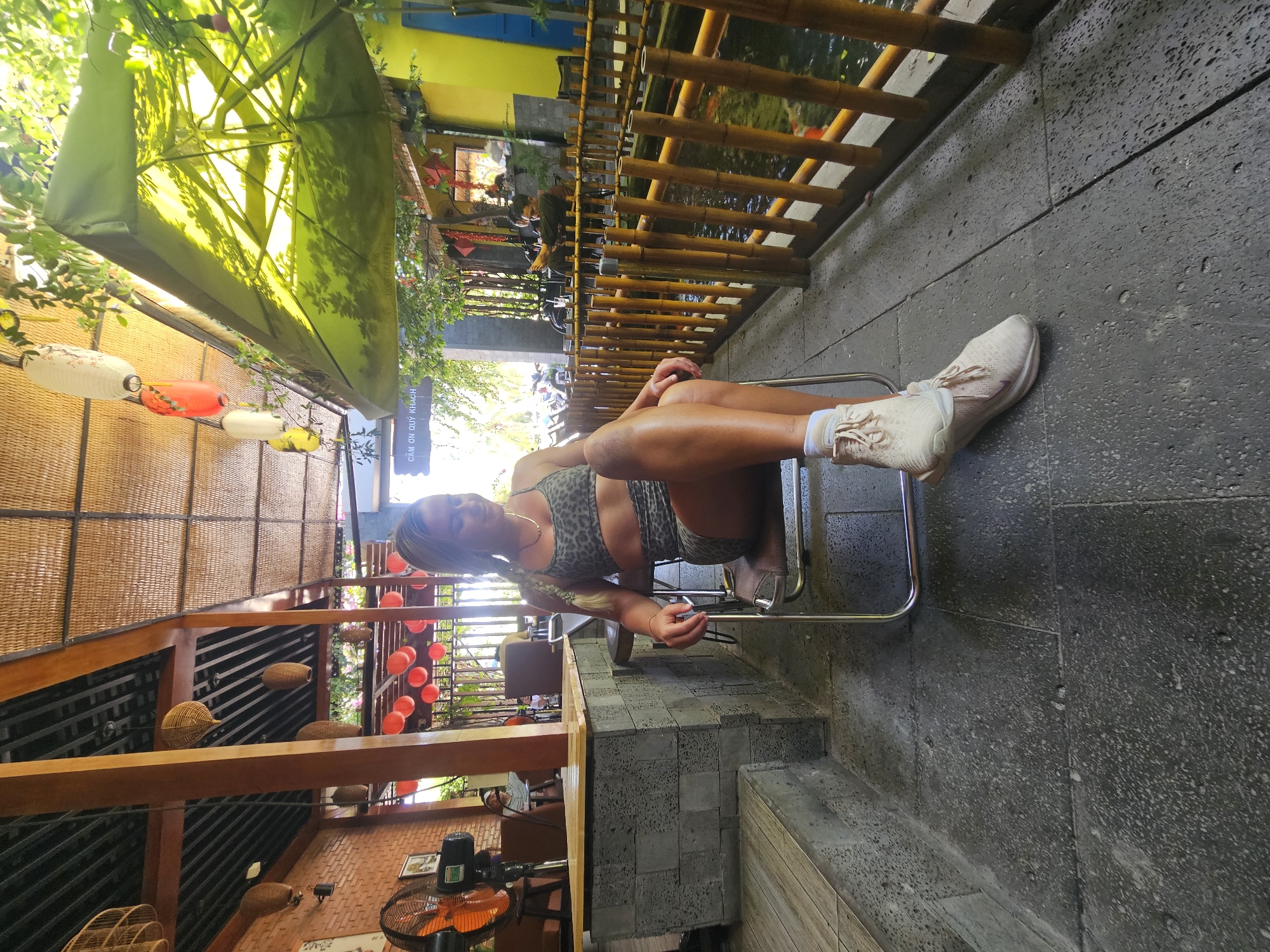 A blonde woman with braided hair is sitting in a metal chair outdoors. She is wearing a leopard-print sports bra and shorts. Her legs are crossed and she is looking down. The background shows a covered outdoor seating area with bamboo fencing, a koi pond, and greenery. There are also paper lanterns hanging from the ceiling. The scene appears to be a cafe or restaurant in Mui Ne, Vietnam.