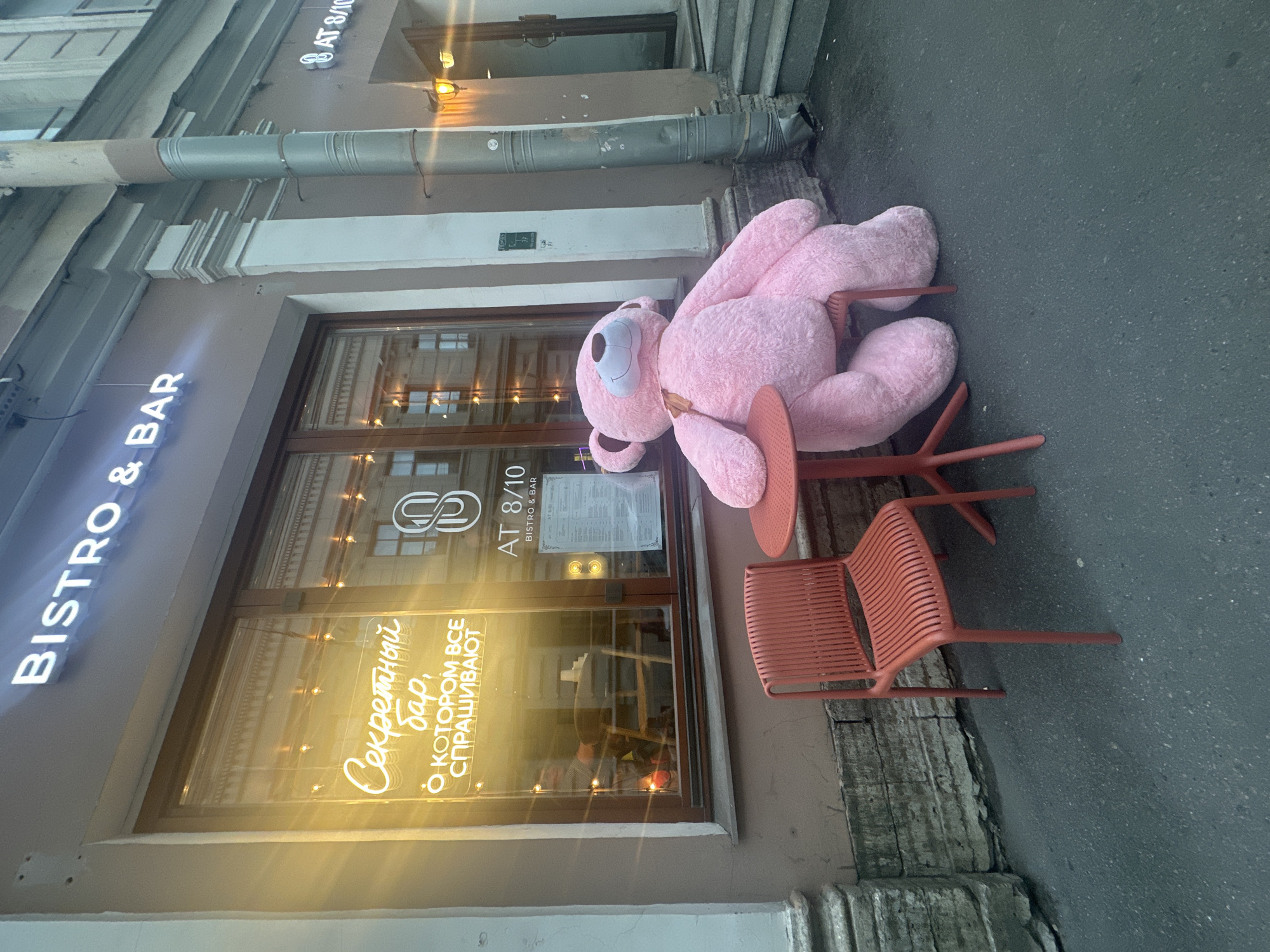The image shows a large pink teddy bear sitting on a chair outside a bistro and bar in what appears to be Saint Petersburg, Russia, based on the Cyrillic signage. The restaurant is named "AT 8/10" and advertises a "secret bar." The bear is seated at a small outdoor table with two chairs. The street appears wet or recently washed.