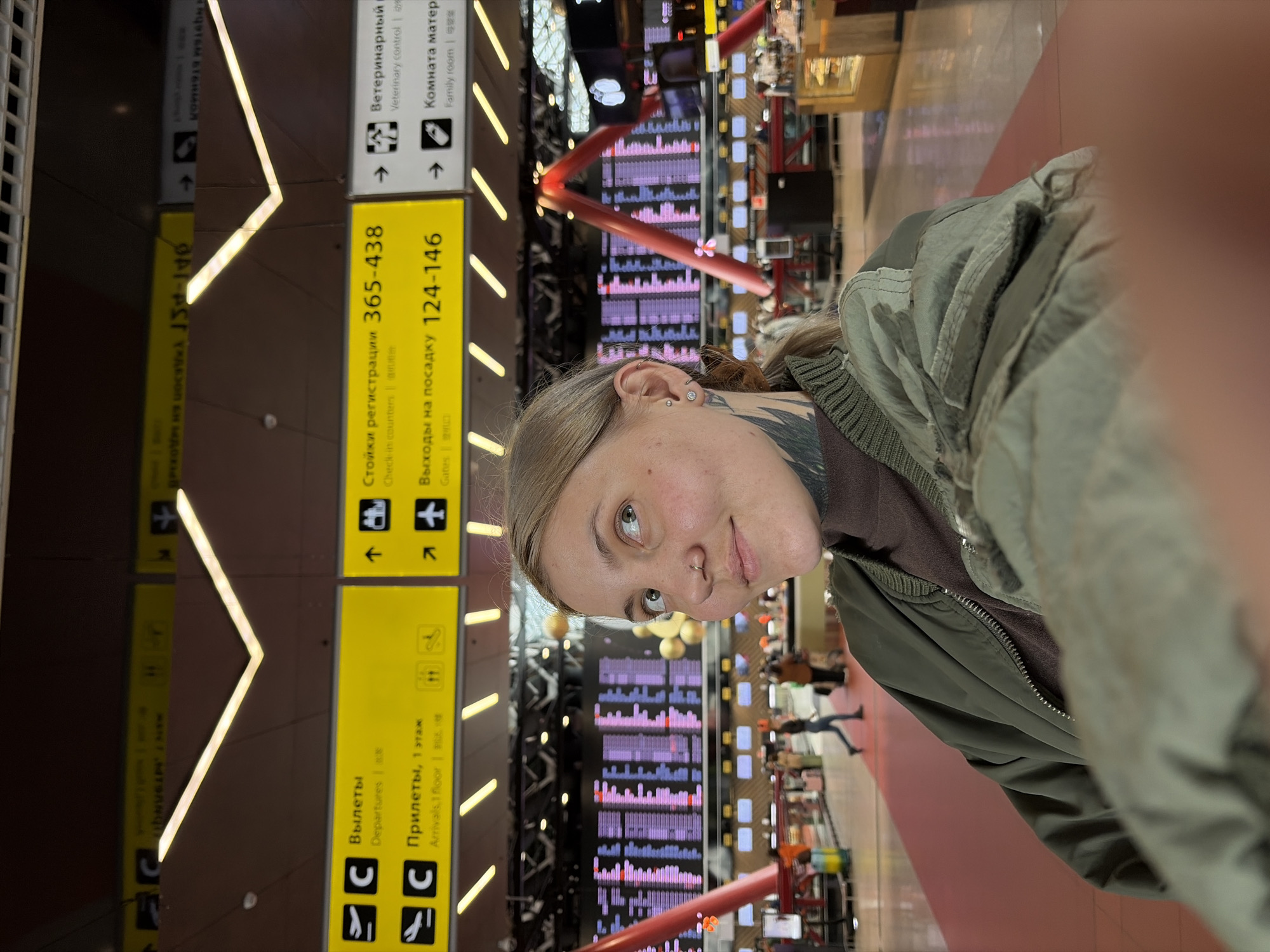 A woman with light hair and a nose ring is in what appears to be an airport terminal. Signage in Russian is visible, indicating check-in counters 365-438 and gates 124-146, along with directions for "Departures" and "Arrivals." An electronic display board showing flight information is behind her. The presence of Russian text suggests a location within Russia. Given that Sheremetyevo International Airport (SVO) is a prominent airport near Khimki, Russia, it is likely that this scene is located in Sheremetyevo.