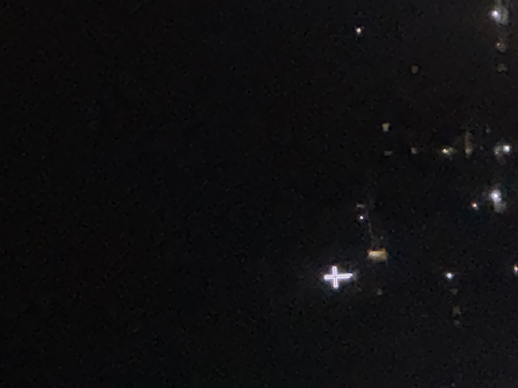 The image appears to be a nighttime scene in a city, potentially Yerevan, Armenia, judging by the scattered lights in the background. There's a prominent plus sign (+) visible in the lower right quadrant of the image. The rest of the image is largely dark with variations in brightness. The overall quality is poor with visible image noise.