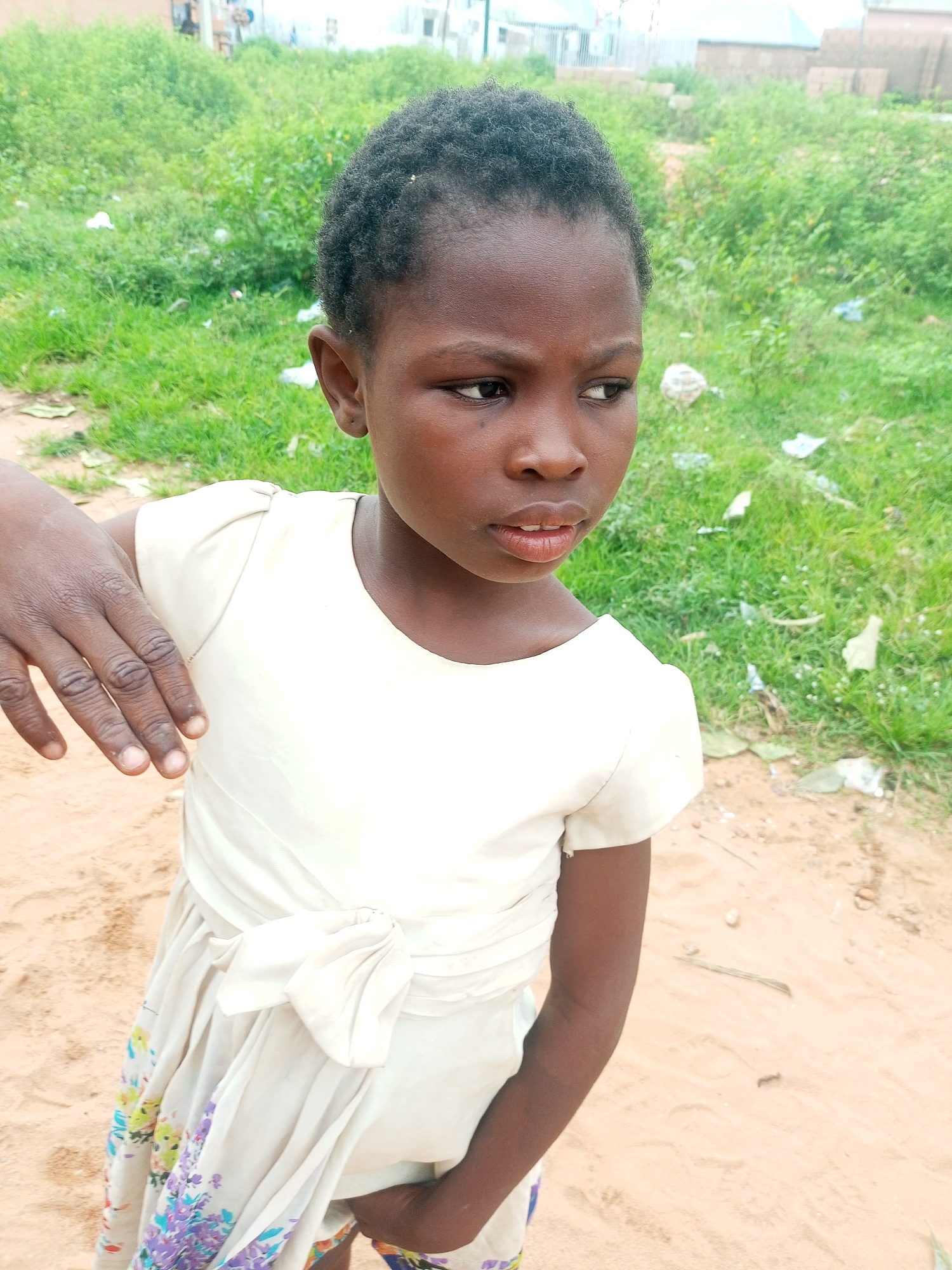 A young girl stands in an outdoor setting. She is wearing a white dress with a bow tied at the waist and a floral pattern at the hem. Her hair is short and dark. She is looking off to the right of the frame with a slightly pensive expression. Her left arm is extended, with her hand open and resting on her chest. The background is a grassy area with some scattered debris, and in the distance, there are buildings and structures. The ground in the foreground is sandy. The lighting suggests it is daytime, and the overall scene appears to be in a rural or semi-urban area in Tempure, Nigeria. There is no visible text.