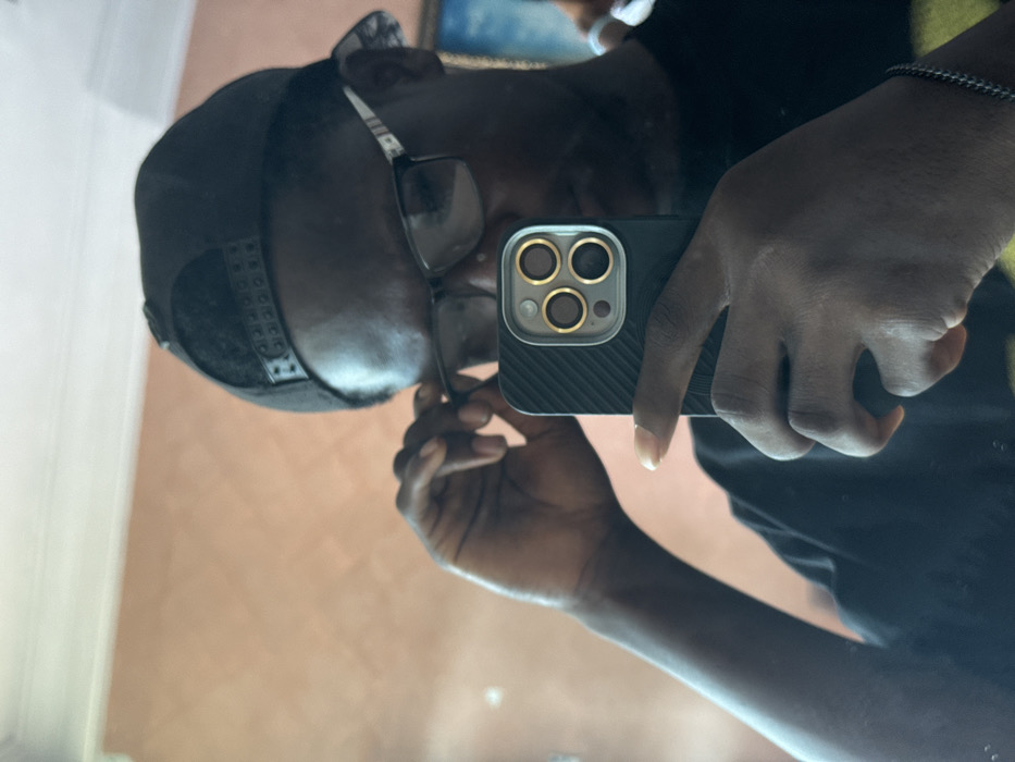 A male individual with dark skin is depicted taking a mirror selfie indoors in Eziobodo, Nigeria. The subject wears a black t-shirt, a black baseball cap reversed, and dark-framed rectangular eyeglasses with silver or white accents on the temples. The subject holds a black smartphone, equipped with a black ribbed case and a visible triple camera array featuring gold lens rings, in their right hand. Their left hand is positioned near the subject's right temple, adjusting the eyeglasses. A thin, silver-colored chain bracelet is present on the subject's left wrist. The background consists of a uniformly painted peach-colored wall. A faint reflection of what appears to be a framed item with a blue hue is visible in the upper right background.
