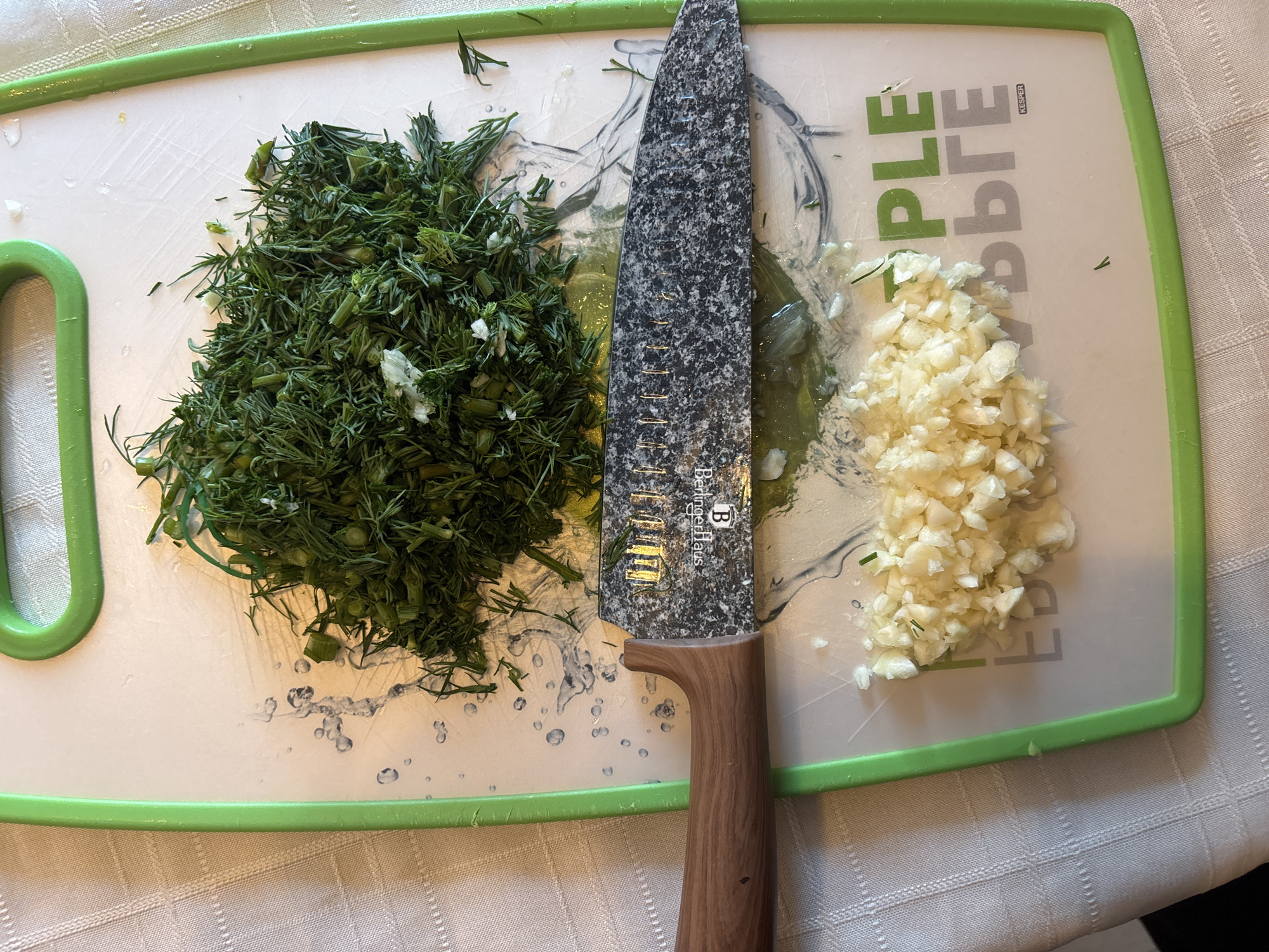 A close-up, overhead shot shows a pile of chopped dill and minced garlic on a white cutting board with green trim. A knife with a gray speckled blade and wooden handle is positioned across the cutting board, dividing the dill from the garlic. Water droplets are scattered on the cutting board. The cutting board rests on a white checkered tablecloth. Text on the cutting board reads "BERLINGERHAUS" and the word "KSPER" is visible near the bottom right.