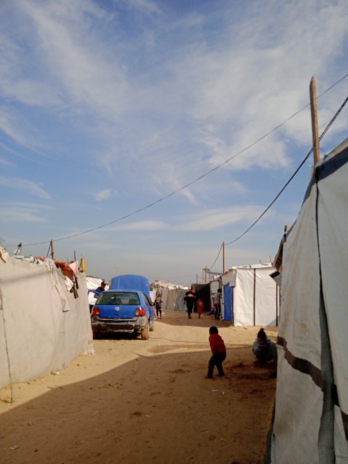 The image depicts a dirt pathway through a dense, informal settlement in Khan Yunis, Palestinian Territory, under a bright blue sky with scattered cirrus clouds. The pathway, composed of light brown, dry earth, recedes into the distance, lined on both sides by numerous makeshift structures constructed from white and blue tarpaulins and other sheeting materials.
On the left side of the path, a blue hatchback vehicle, possibly a Volkswagen Golf, is parked with its hood open and its rear bumper missing. An individual in dark clothing is partially visible near the open hood, looking into the engine compartment. Further down the path on the left, laundry or fabric items are draped over the exterior of one of the shelters.
In the immediate foreground, a child wearing a red shirt stands with their back partially to the camera, facing left. To their right and slightly behind, another individual is crouched low to the ground. Further down the path, beyond the blue car, a group of three or more figures, including adults and children, walk away from the camera.
Overhead, black electrical or communication wires traverse the scene, suspended between wooden poles visible on both sides of the path, connecting to the various shelters. The sun casts distinct shadows, indicating bright daylight conditions.