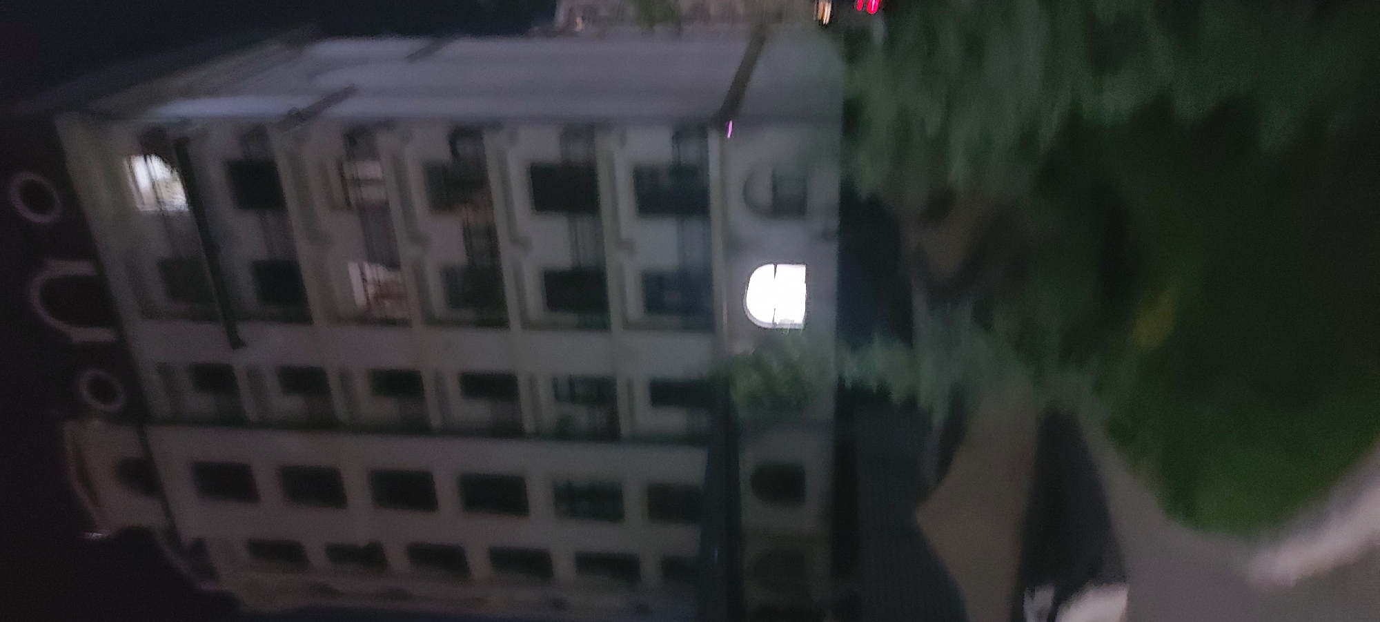 Here is a description of the image.
The image shows the architecture and urban environment of Tashkent, Uzbekistan. A multi-story building with classical architectural features is visible, characterized by repeating window patterns and decorative elements. The building is illuminated, with light emanating from some windows. Vegetation, including trees or shrubs, is present on the right side of the frame, partially obscuring the building. The scene appears to be taken at night, with subdued lighting conditions. A sidewalk or paved area is visible in the foreground.