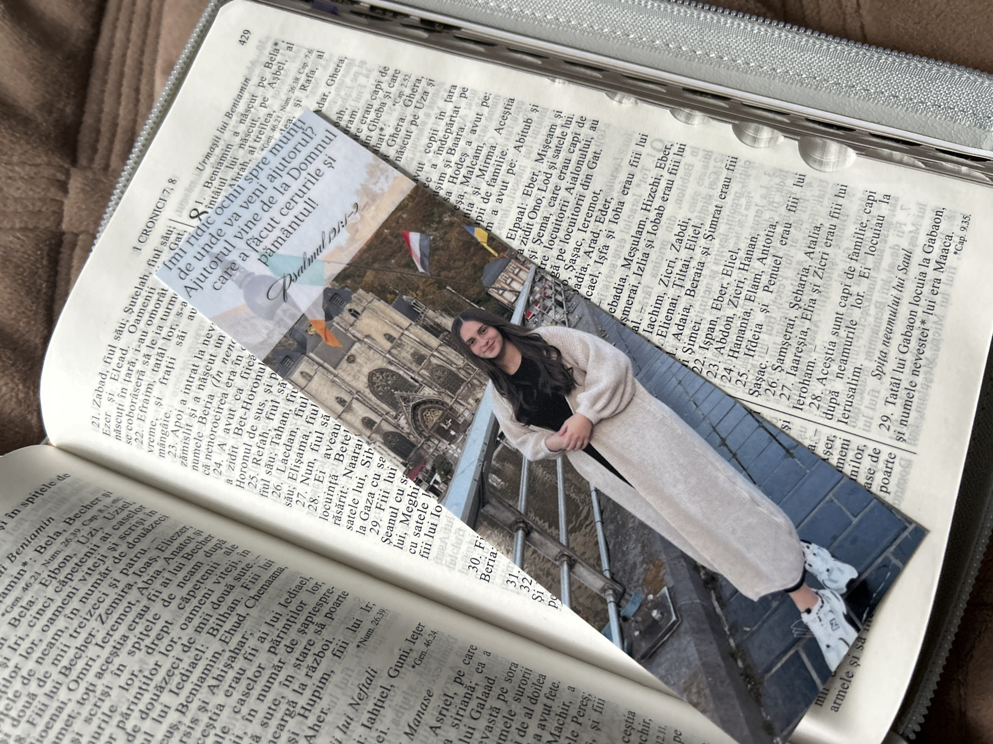 The image shows an open Bible with a bookmark featuring a photograph of a young woman. The Bible is open to a page in 1 Chronicles, with text in Romanian. The bookmark's photograph is placed diagonally across the open pages. The bookmark also has text that reads "Psalm 121:1-2" and "Ajutorul vine de la Domnul care a făcut cerurile și pământul!!" which translates to "Help comes from the Lord who made the heavens and the earth!!". The background of the bookmark appears to be a building, possibly a church or historical structure, with flags visible. The setting is indoors, on a surface that appears to be a table or desk. The image is taken from a slightly elevated angle.