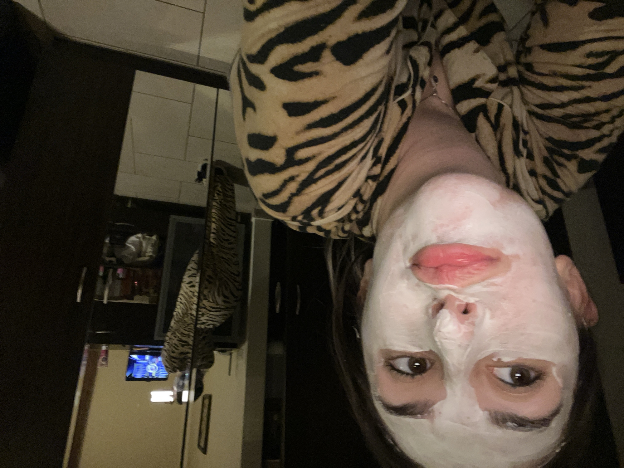 The image is an indoor selfie of a person wearing a tiger-striped shirt and a white face mask. Behind the person, there's a glimpse of a room with dark furniture, what looks like a television screen reflecting in a dark surface, and a tiled wall. The photograph appears to be taken inside a bathroom or a similar space, but there is no information to indicate the location in the city of Sofia, Bulgaria.