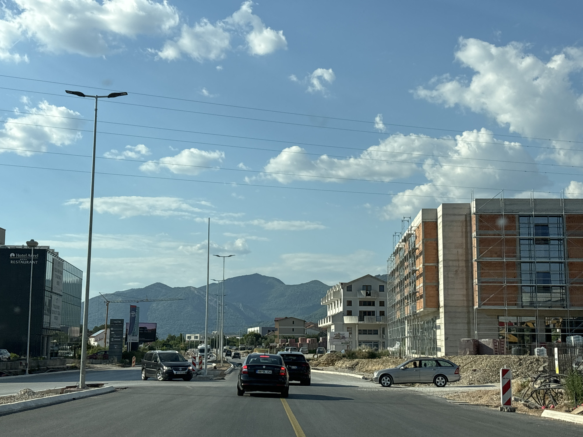This media file captures a view from a vehicle driving on a road in Radanovići, Montenegro. The scene depicts a developing urban area with ongoing construction, evidenced by buildings covered in scaffolding and piles of building materials. On the left, a modern, dark-glass building with signage for "Hotel Aurel" and "RESTAURANT" stands prominently. In the distance, rolling, tree-covered mountains form a natural backdrop under a bright blue sky dotted with fluffy white clouds.
Several vehicles are visible on the road: a black Volkswagen SUV in the foreground, followed by other cars and a van further down the street. A silver car is parked on the right side of the road near a construction site. Two tall streetlights are positioned along the road, and power lines crisscross the sky.
The time of day appears to be late afternoon or early evening, suggested by the warm light and long shadows. The weather is clear and sunny, with a few clouds contributing to a picturesque sky. The overall impression is one of progress and growth in a scenic, mountainous region.
Visible text includes "Hotel Aurel", "RESTAURANT", and various signs related to businesses and potentially advertisements, such as one for "TOYOTA". License plates are also visible on some vehicles, though the text is difficult to read clearly.