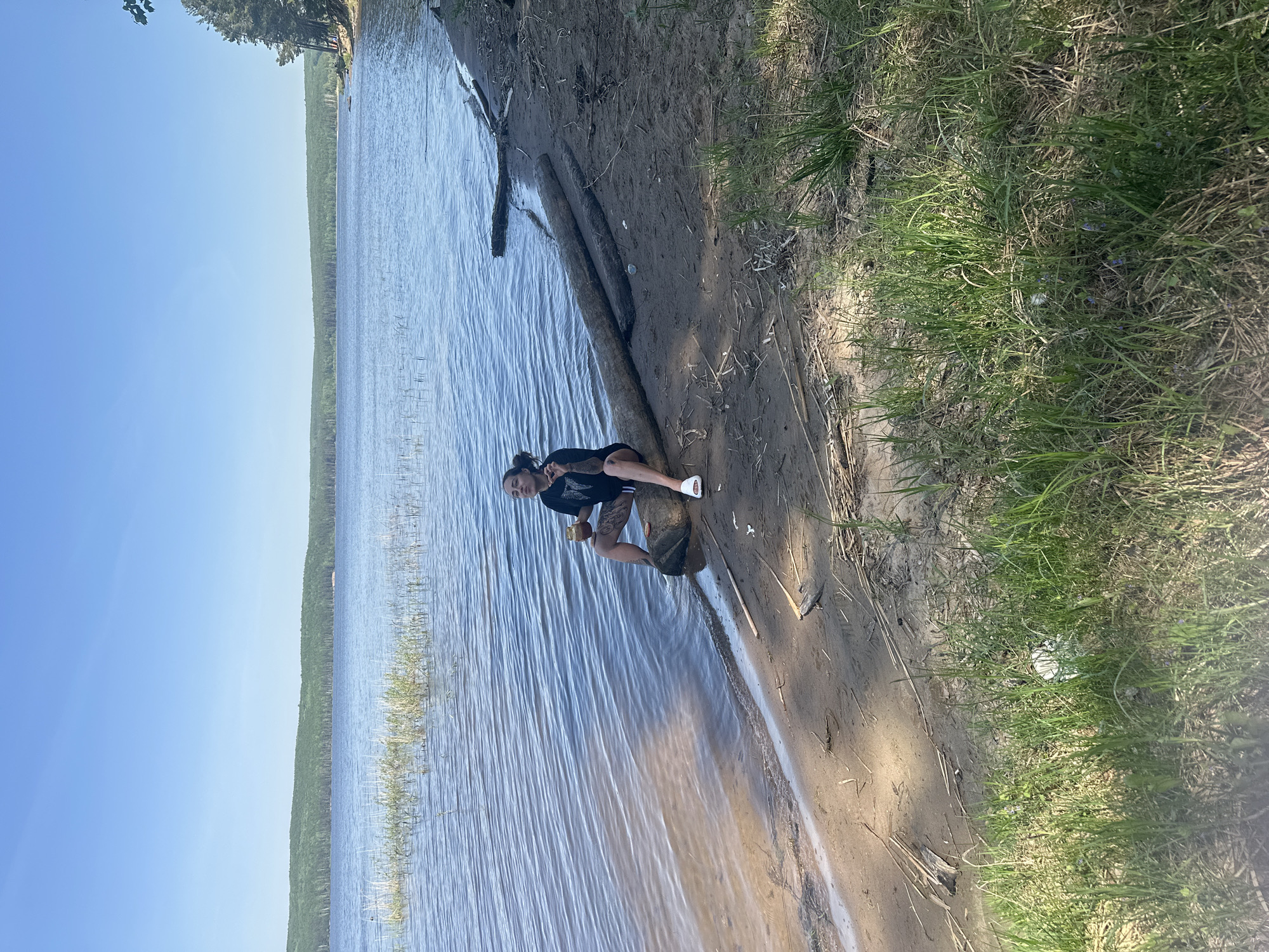 A woman sits on a log on the shore of a body of water, potentially a lake or a wide river. The water stretches towards the horizon under a clear sky. The shoreline consists of sand and grassy vegetation. The location is likely near Chaikovsky, Russia. She is holding an item in her hand and appears to be at peace.