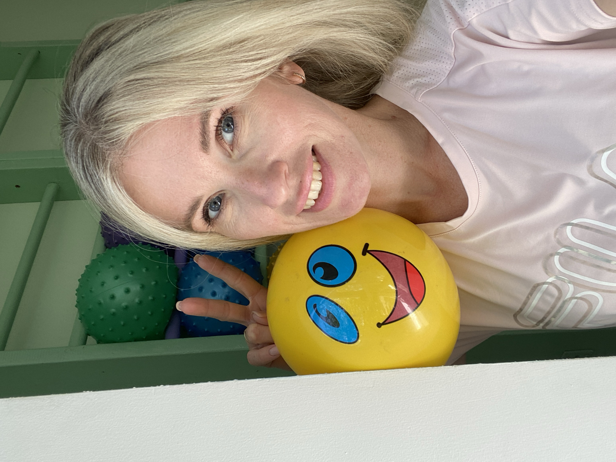 A blonde woman is taking a selfie while holding a yellow smiley face balloon. She is wearing a pink t-shirt and has blue eyes. Behind her, there are several colorful textured balls. The background appears to be a room with green shelving and possibly playground equipment. The lighting suggests it is daytime. The location is described as Unknown, Unknown.