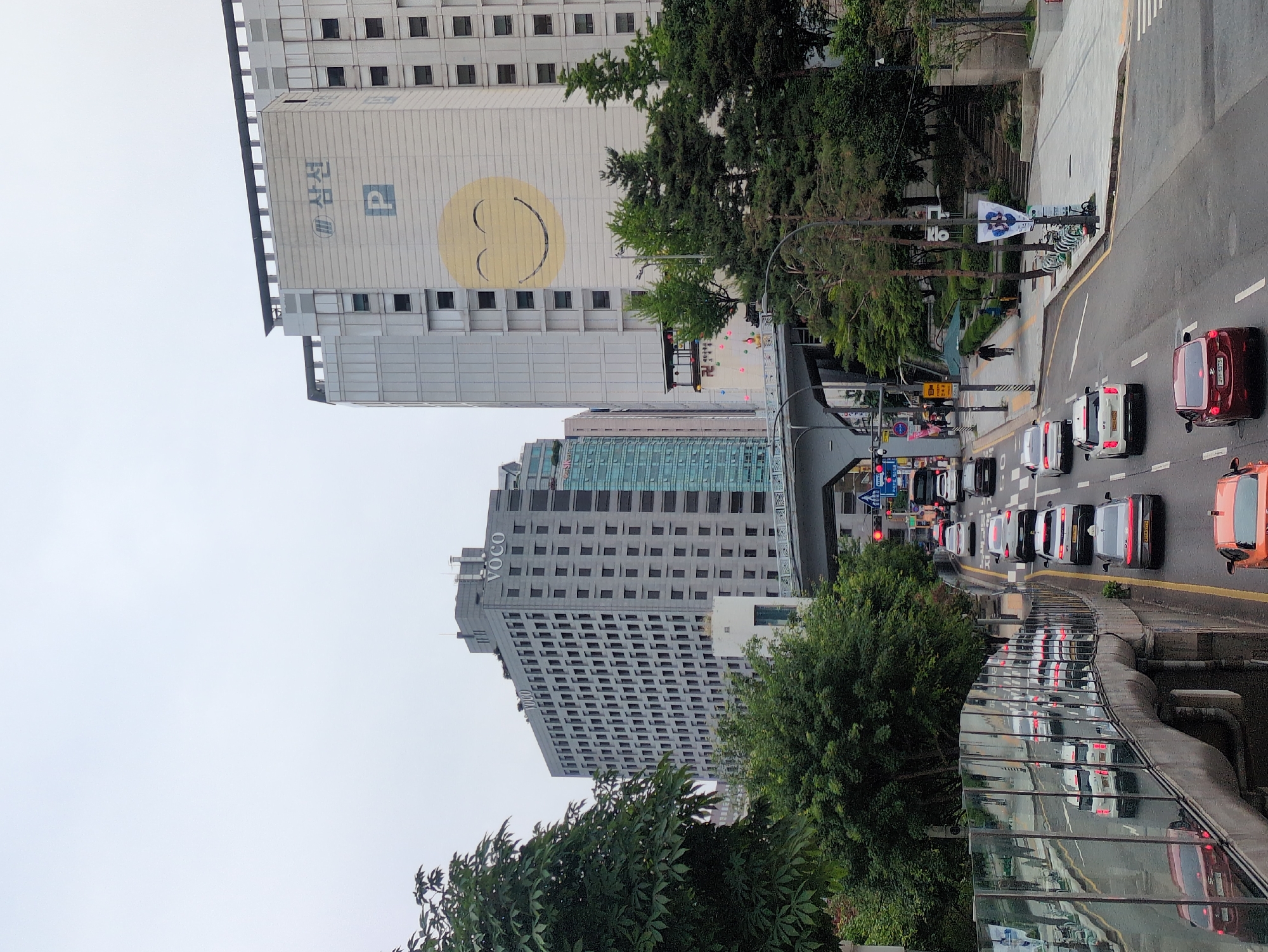 This image depicts an urban street scene in Seoul, South Korea, specifically located in the Gangnam district, identifiable by the Voco Seoul Gangnam hotel (marked "VOCO") and the prominent building displaying "삼성" (Samseong) with a large yellow smiley face mural. The scene is viewed from an elevated perspective on an overcast day.
In the foreground on the left, a curving, reflective glass sound barrier partially obscures a lower road segment, mirroring the vehicles and street environment. Dense green foliage of trees extends from the left margin.
The central midground is dominated by a multi-lane asphalt road with numerous automobiles, primarily sedans and SUVs, adhering to right-hand traffic. Vehicles of various colors including white, grey, red, black, and orange are visible, some appearing stationary and others in motion. A red traffic light is active further down the road. A grey concrete elevated pedestrian bridge or overpass spans diagonally across the midground, its understructure visible.
To the right of the roadway, a concrete sidewalk is present, populated by a few pedestrians. A row of shared bicycles is parked along the sidewalk's edge. Streetlights, a blue and white banner with Korean text, and signage are visible along the pedestrian pathway.
The background features prominent high-rise buildings under a uniformly light grey, overcast sky. On the right, a tall, light-colored building, identified as a Samseong facility, displays a large yellow circular smiley face mural with a simple curved mouth and closed eyes. Above this, the Korean text "삼성" (Samseong) and a blue 'P' (for parking) are affixed to the facade. Below the smiley face, a banner with a red Buddhist swastika symbol is partially visible. To the left, a dark grey building, identified as the Voco Seoul Gangnam hotel by multiple "VOCO" signs on its roofline and upper facade, features a grid pattern of numerous dark, rectangular windows. Further left, a taller building with a modern blue-tinted glass facade is partially visible. Abundant green trees and foliage are interspersed throughout the scene, particularly along the road's edges and in front of the buildings.
