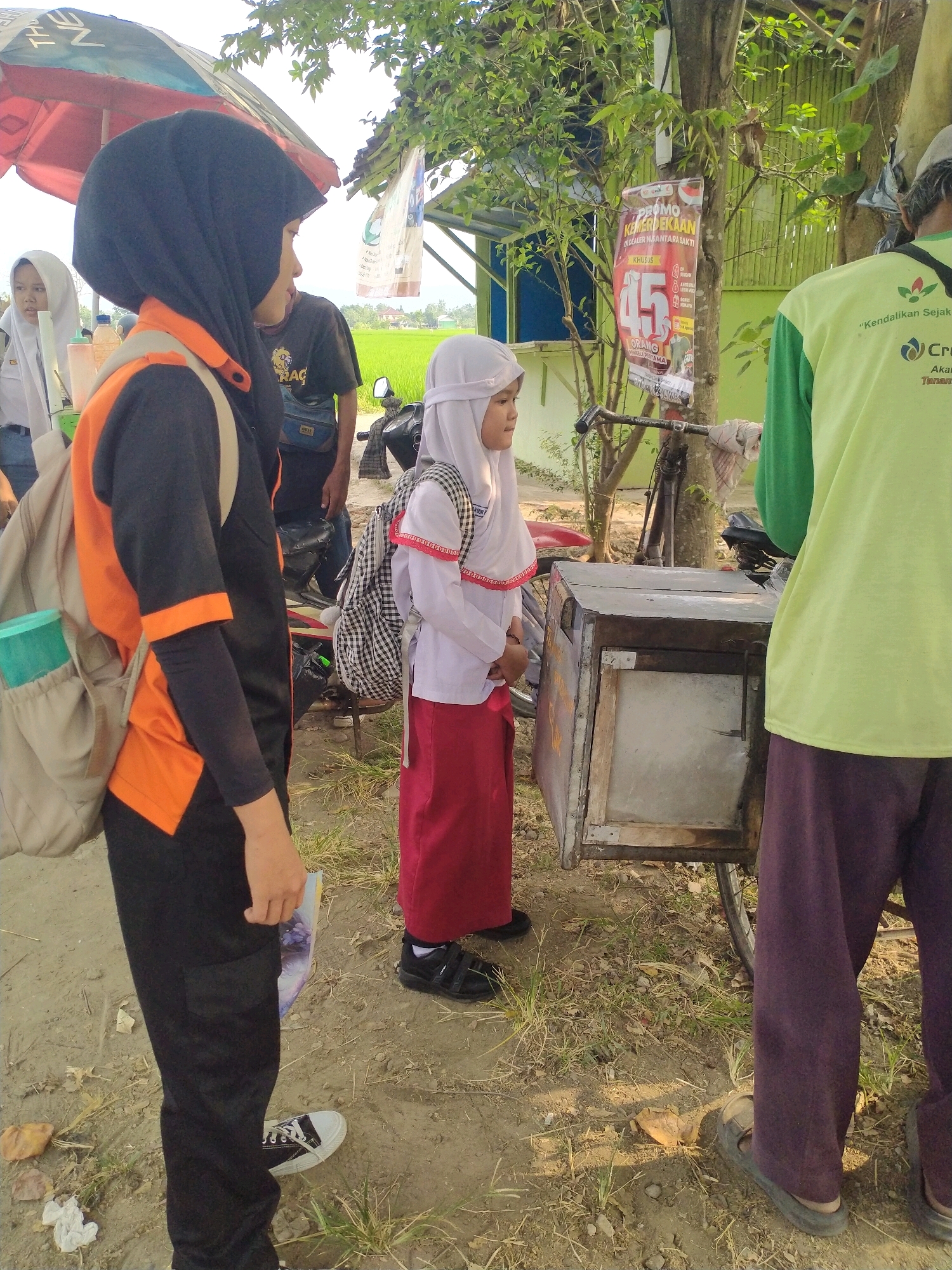 In Begajah, Indonesia, a group of four people are gathered near a roadside stall. The foreground features a person wearing a black hijab and an orange and black uniform, with a beige backpack slung over their shoulder. To their right, a child in a white hijab and a white and red school uniform stands looking at something in the stall. Behind them, a person wearing a light green shirt and dark purple pants is partially visible, facing away from the camera. To the far left, another young person in a white hijab and grey uniform is also present. The environment includes a dirt ground, dry grass, trees, and a red and white umbrella providing shade. A bicycle is parked next to the stall, which appears to be a cart with a wooden and metal construction. A promotional poster with Indonesian text and the number "45" is attached to a tree. The background shows a rice field under a bright sky.