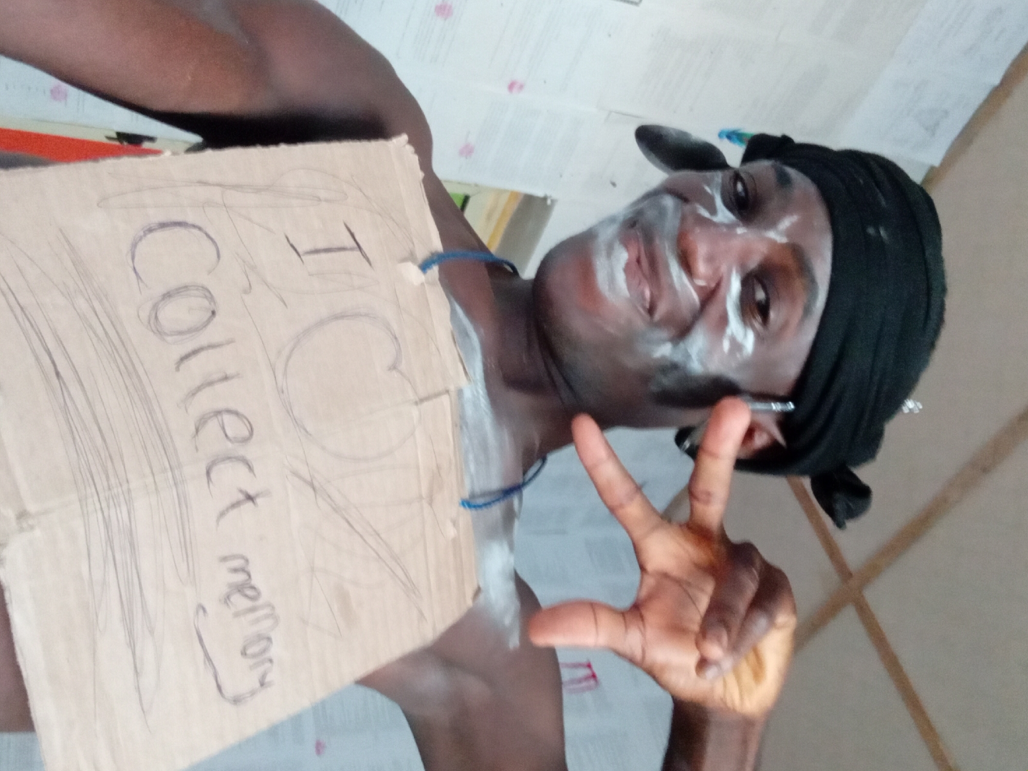 A man is standing inside, holding a cardboard sign that reads, "I Collect money." He has white markings on his face and neck, and wears a black head wrap. He is making a hand gesture with his right hand. The location appears to be an interior space with neutral-colored walls. The city is Unknown, Unknown.