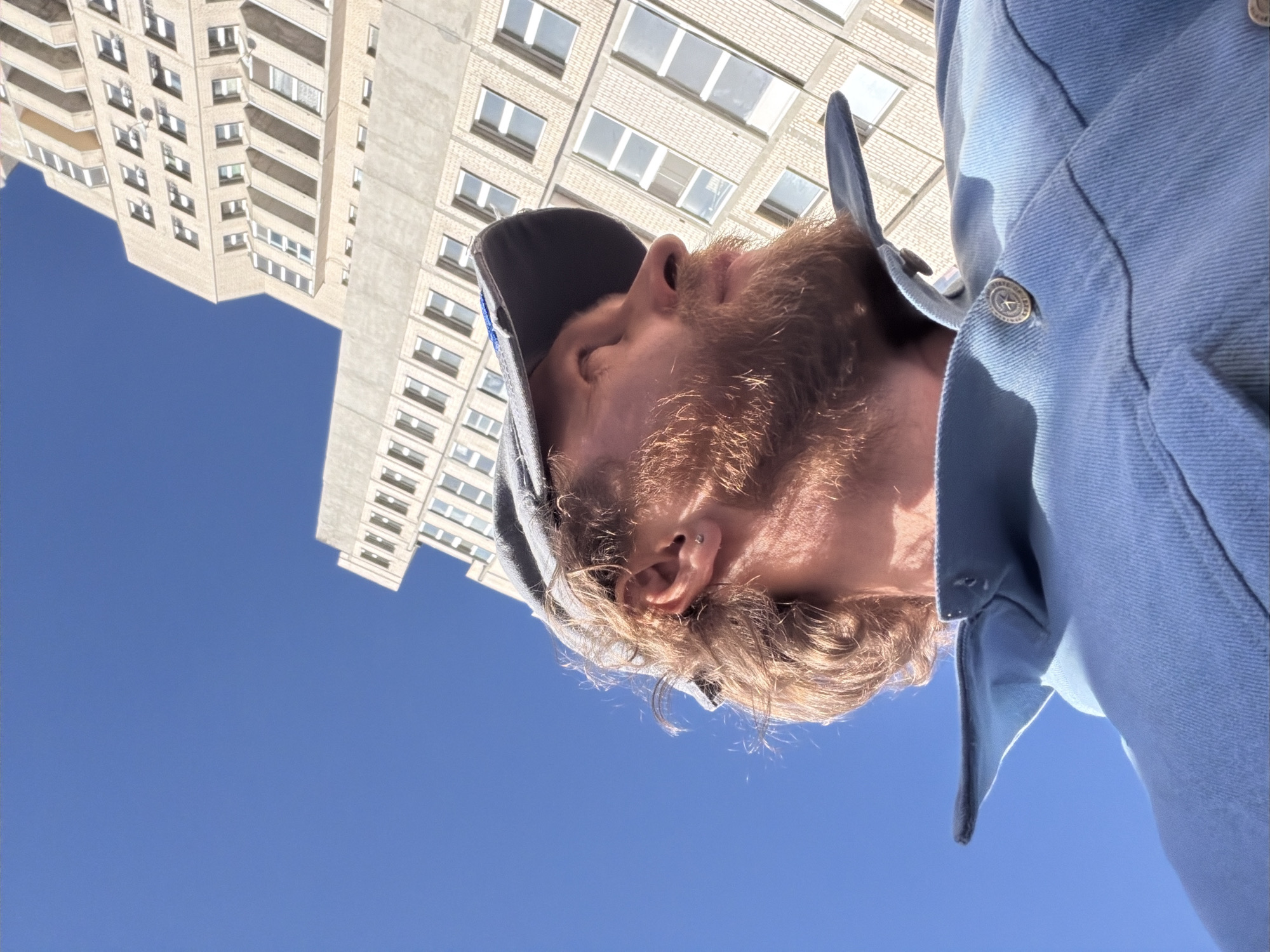 The media file presents an upward-facing, rotated close-up shot of a man's head and shoulder against a bright blue sky and a tall building. The subject is a man with a prominent reddish-brown beard and mustache, and blondish hair visible around his ears. He is wearing a dark baseball cap, partially visible on his head, and a light blue denim-style jacket. A silver button with a star design and faint, unreadable text is visible on his jacket. He also has a tragus piercing in his visible right ear. The man's face is seen from the chin up, with his eyes appearing to gaze upwards.
In the background, a large, multi-story building constructed from light-colored brick, featuring numerous windows and balconies, rises towards the sky. The architecture suggests a residential or commercial complex, typical of a large city. The clear, vibrant blue sky indicates a sunny day, likely late morning or early afternoon, with no clouds. The scene captures an urban environment in Saint Petersburg, Russia. The unusual, low-angle perspective creates a dynamic and somewhat abstract composition. No specific activity beyond looking up is clearly identifiable, and due to the partial view of the face, no distinct emotions are discernible.