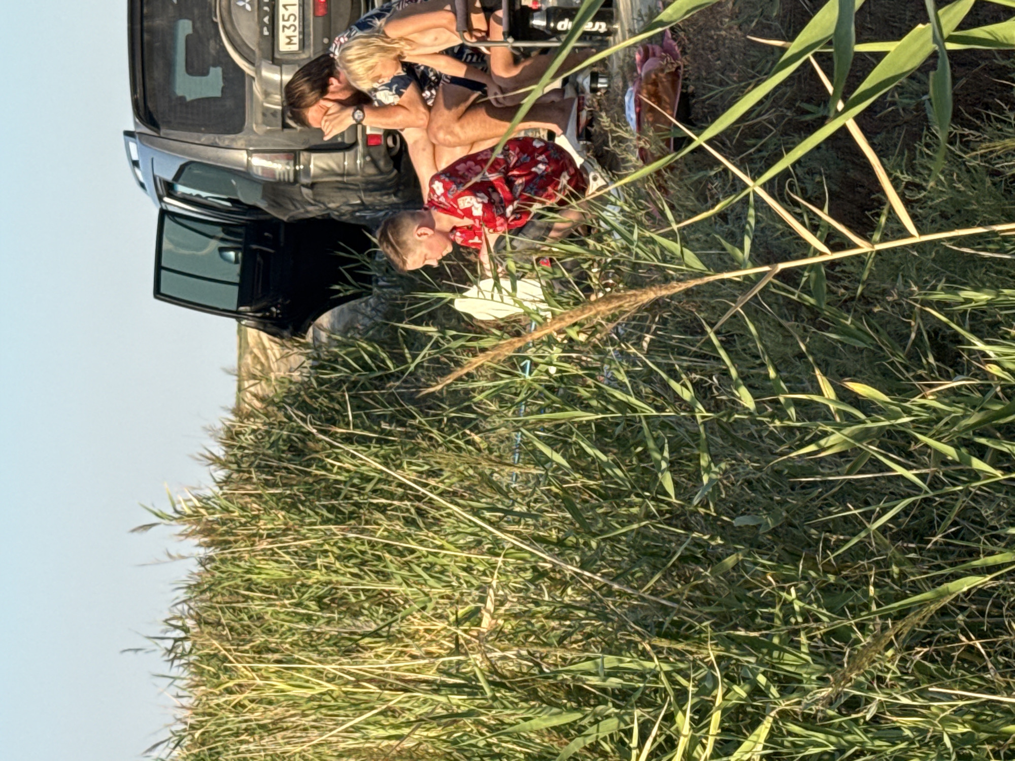In a grassy area with tall reeds, a dark-colored SUV is parked with its doors open. Three individuals are visible: a man with dark hair, wearing a patterned shirt, sits with his hands near his face. Next to him, a young boy in a red, patterned shirt is crouching. To the right of the man, a fair-haired child is seated on a chair. The ground is covered in dry grass and some foliage, with a spare tire on the back of the SUV and a license plate visible. The sky is clear and blue.